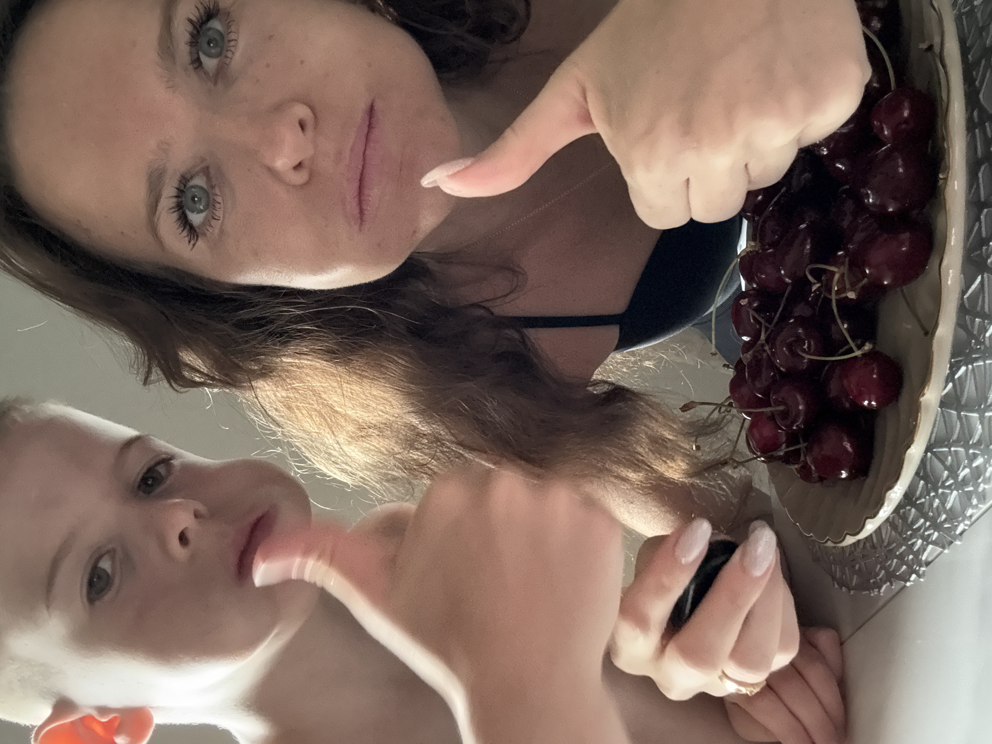 In Dolgoprudny, Russia, a close-up, low-angle shot shows a woman and a young boy. The boy is to the left, with his thumb up. He has light brown hair, fair skin, and a slightly pensive expression. The woman is to the right, also with her thumb up, and appears to be looking at the camera. She has long, dark brown hair and fair skin with freckles. Both are close to the camera, with a plate of dark red cherries in the foreground. The woman is wearing a dark-colored top. The background is indistinct, suggesting an indoor setting.