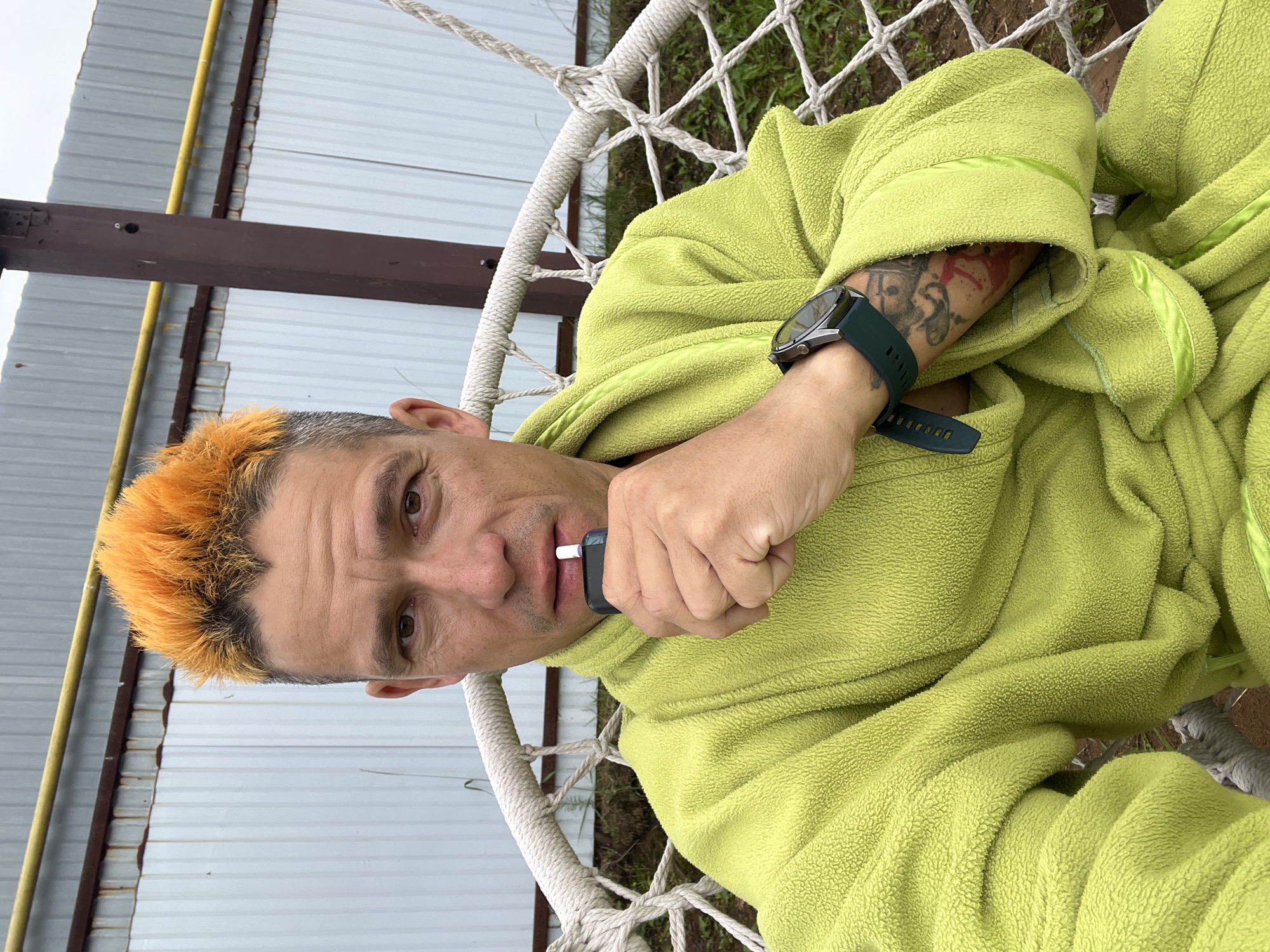 In a selfie taken outdoors, a man with bright orange, spiked hair sits in a hammock chair. He is wearing a fluffy, lime-green bathrobe. The man is holding a dark-colored electronic cigarette device to his mouth. Visible on his left wrist is a dark-colored smartwatch with a teal band. Tattoos are visible on his left arm, peeking out from the sleeve of his bathrobe. In the background, a corrugated metal wall with horizontal yellow piping runs across the frame. A brown metal pole, likely part of a structure, stands to the right of the man. The hammock chair is constructed with thick white rope. The ground beneath the chair is covered with grass and dirt. The scene appears to be in a yard or outdoor recreational area, possibly in Chaikovsky, Russia, given the man's apparent nationality and the utilitarian construction often seen in such areas.