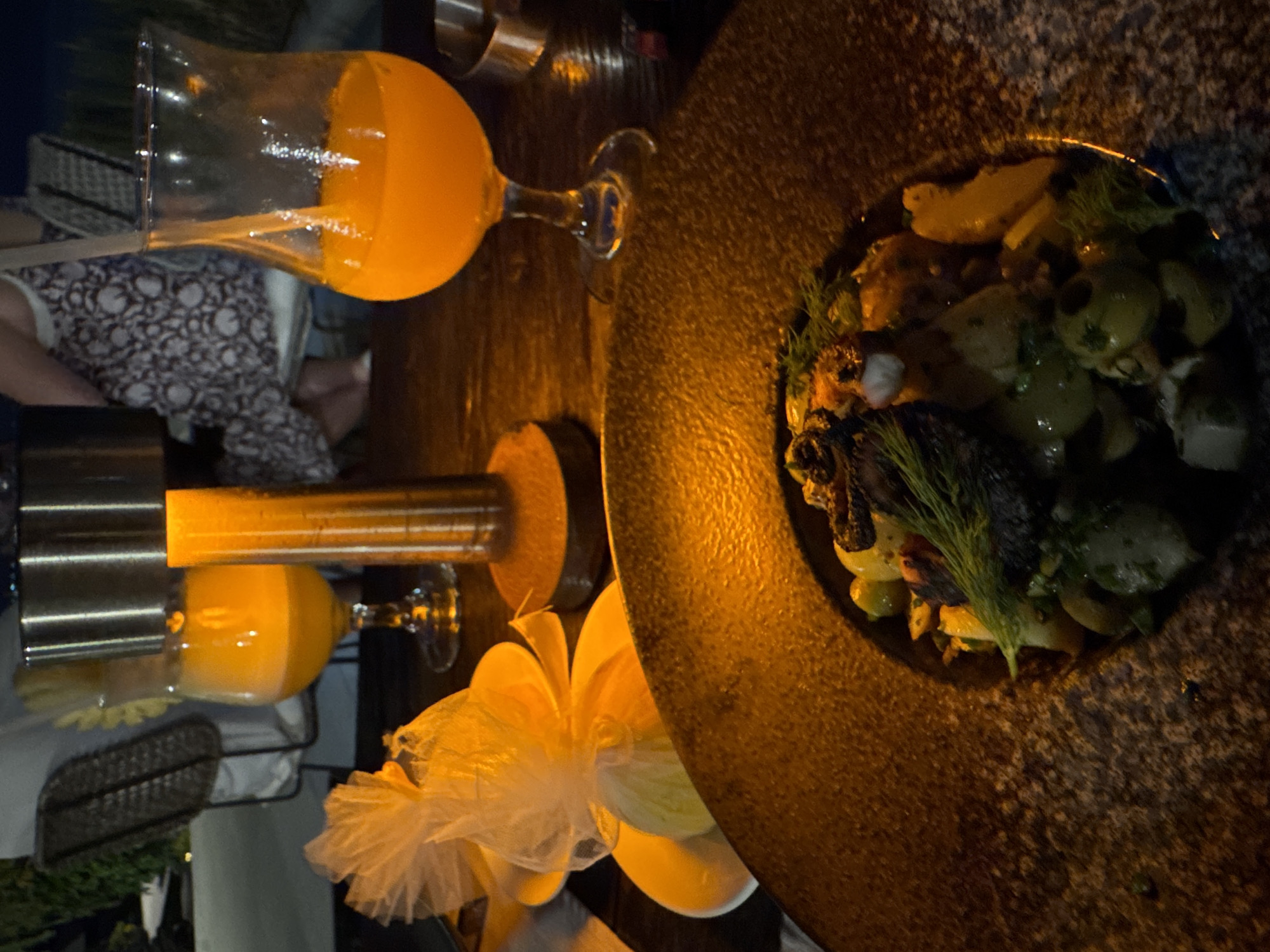The image captures a dimly lit dining scene at night, likely in Kuta Selatan, Indonesia, creating an intimate and warm ambiance. The primary subjects are a meticulously plated meal and two glasses containing orange beverages, arranged on a dark, textured table.
On the right, a large, dark, and somewhat rough-textured plate holds an elaborate dish. The meal appears to consist of various ingredients, including what looks like seafood, possibly octopus or calamari, alongside green components such as olives or capers, and fresh green herbs like dill or fennel fronds. The dish is illuminated by a warm, orange glow, highlighting its textures and colors.
To the left, a clear stemmed glass is filled with a bright orange liquid, resembling juice or a cocktail, with visible bubbles, suggesting it might be freshly poured or carbonated. Further left, another similar stemmed glass, also containing an orange drink, is positioned directly beneath a tall, cylindrical metal dispenser or cooler. This dispenser emits a strong, warm orange light, which serves as a central light source for that part of the table.
In the mid-ground, a delicate, light-colored object, possibly a ruffled napkin or decorative element made of sheer fabric, adds a soft contrast to the dark table and strong lighting. A person is partially visible in the blurred background, seated, wearing patterned clothing, adding to the dining atmosphere. The overall activity depicted is dining and enjoying drinks in a sophisticated setting during the evening. No visible text is present in the image.