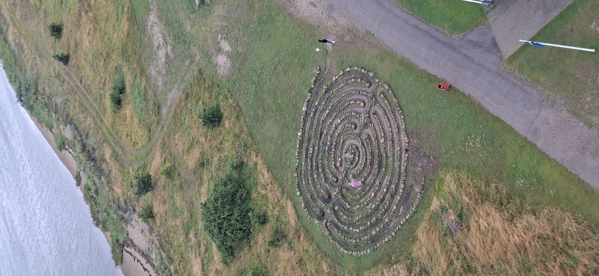 In the city of Sääre, Estonia, an aerial view captures a circular stone labyrinth situated on a grassy hill. The labyrinth's intricate design is formed by a winding path of stones, encircling a central area. A person dressed in pink is visible within the labyrinth, walking along the path. To the right of the labyrinth, a paved path leads away, and two other individuals are present on this path. One person, wearing a dark jacket and white shoes, stands near the labyrinth, while another, dressed in red, is further down the path. The surrounding environment is a mix of green grass, dry, tall grasses, and sparse bushes. In the distance, a body of water borders the landscape, with a sandy shoreline. A flagpole with a blue and white flag is positioned in the lower right corner of the image, adjacent to a paved area.