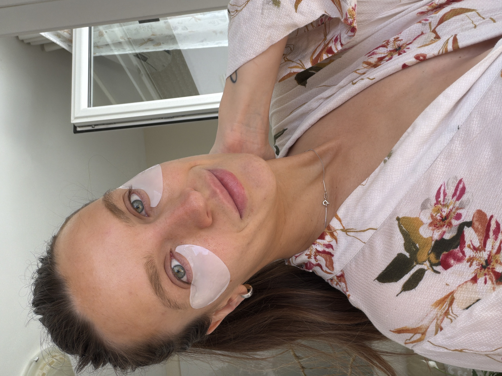 A woman with green/hazel eyes, long brown hair, and defined eyebrows is captured in a close-up portrait, oriented sideways as if reclining. She looks directly at the camera with a subtle smile. She wears white, translucent eye patches beneath both eyes, suggesting a self-care routine. Her attire is a light-colored, textured robe or top adorned with a vibrant floral pattern featuring red, orange, and green blossoms and leaves.
A thin silver necklace with a small, circular charm hangs around her neck, and a silver hoop earring is visible in her left ear. Her right arm is raised, with her hand resting behind her head, revealing a small, dark, indistinct tattoo on her inner forearm. The background features a simple, light-colored wall and a white window frame with glass panes. The window reflects bright, natural light, indicating it is daytime. The overall lighting is soft and even, highlighting her features and the texture of her robe. No text is visible in the image. The setting appears to be an indoor or covered outdoor space, possibly in Budva, Montenegro.