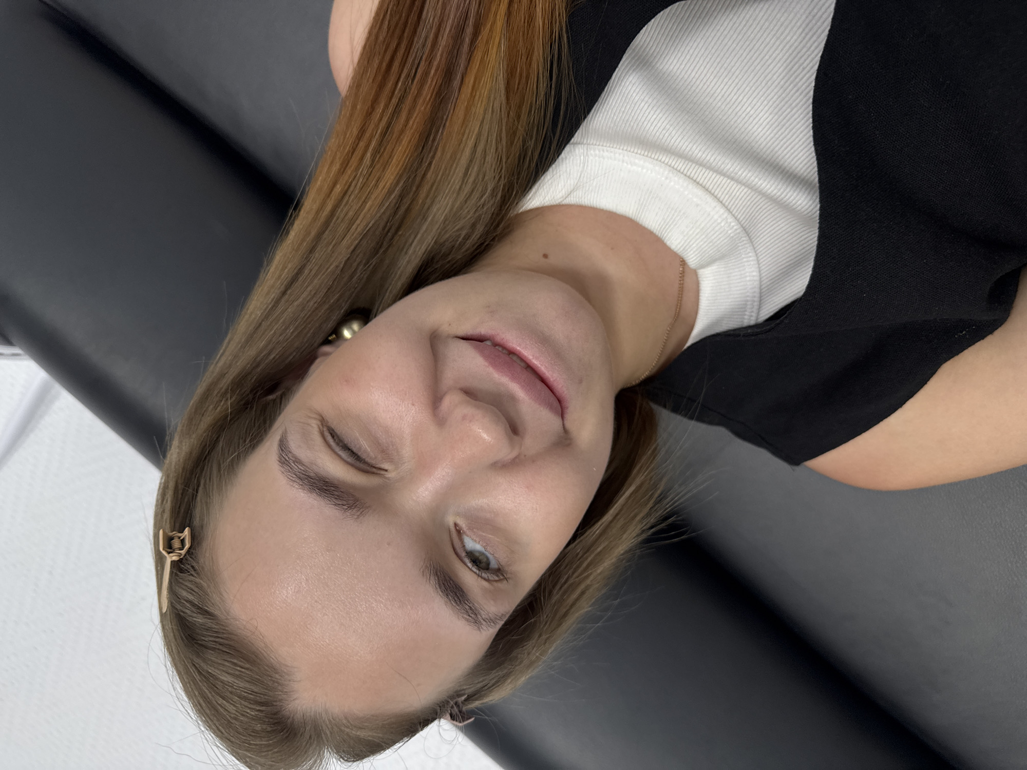 A woman with long, reddish-brown hair and fair skin is lying down on a black surface, possibly a medical examination table. She is wearing a white shirt under a black top. Her right eye is closed in a wink, and she is smiling slightly. A gold-colored hair clip is visible in her hair on the left side of the image. The background shows a white wall and a glimpse of what may be a window. The location is Chaikovsky, Russia.