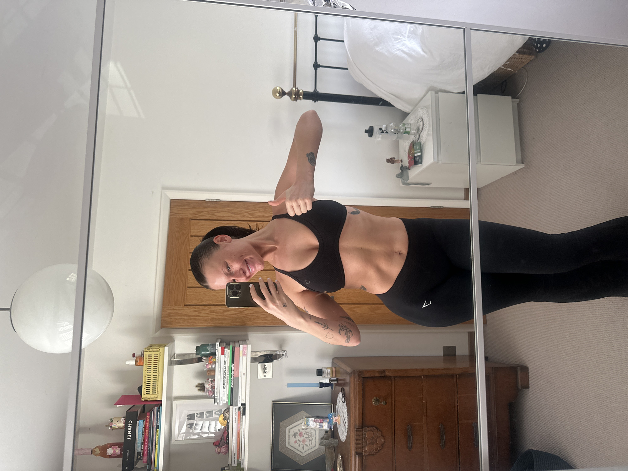 A female individual, appearing in her 30s or 40s, is captured in a full-body mirror selfie within a residential interior in Wealden, United Kingdom. She is dressed in a black sports bra and black athletic leggings, identifiable by a white Gymshark logo on the left hip. Her dark hair is pulled back, and she exhibits a smile. Tattoos featuring botanical motifs are visible on her forearms, with a smaller star-like tattoo on her left wrist. She holds a dark-colored smartphone in her right hand, positioned to capture the reflection, and extends her left arm with a "thumbs up" gesture.
The reflected background reveals a domestic space. To the subject's left, a wooden dresser supports a framed hexagonal piece of art, a light blue tapered candle in a dark holder, and two small amber bottles, likely essential oils, along with other small decorative items. Above this, two white floating shelves are laden with numerous books, including one with "Wealden" visible (reversed) on its spine, a pink Hello Kitty branded bottle, a yellow plastic crate, and small potted succulents. A light brown wooden door is partially visible beyond these furnishings.
To the subject's right, a bed with white bedding and a black metal headboard adorned with brass finials is present. A white two-drawer nightstand stands beside the bed, holding a clear plastic water bottle with a black lid, small figurines, and a white crocheted doily. The floor is covered with a light-colored carpet. A large, spherical white ceiling light fixture is visible at the top of the image.