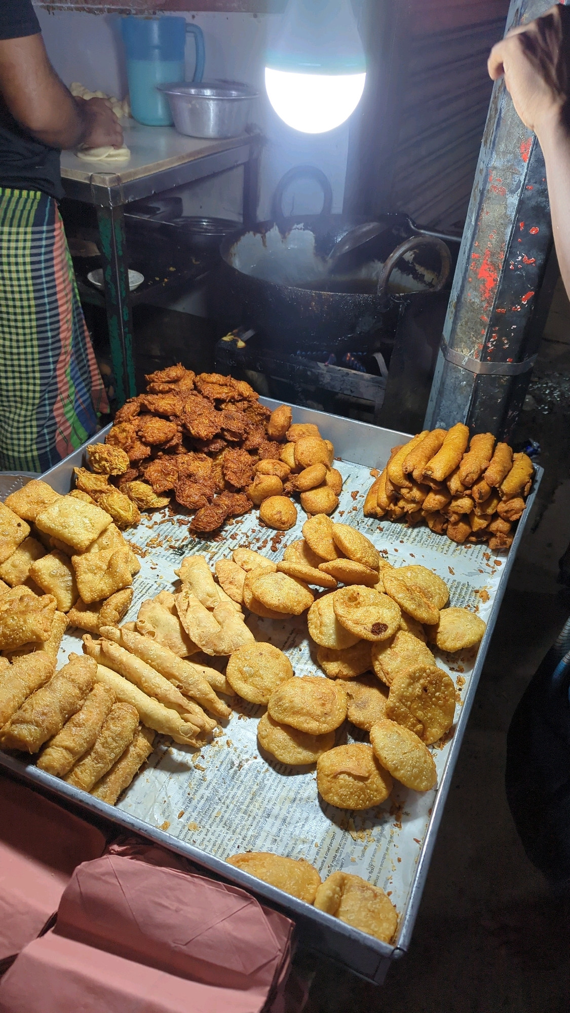 A street food stall in Dhaka, Bangladesh, displays a variety of fried snacks. The snacks are arranged on a metal tray lined with newspaper. In the foreground, there are several types of fried items, including flat, square-shaped fritters, elongated rolls, and round, puffy snacks. Behind these, a large pile of dark brown, irregularly shaped fried items is visible. To the right, another stack of golden-brown, cylindrical fried snacks is prominent. The background suggests a bustling environment, with a person preparing dough on a stainless steel table, a blue pitcher, and a metal bowl. A large wok with hot oil is actively being used for frying, illuminated by a bright LED bulb. The overall scene depicts a typical street food vendor in Dhaka, Bangladesh.