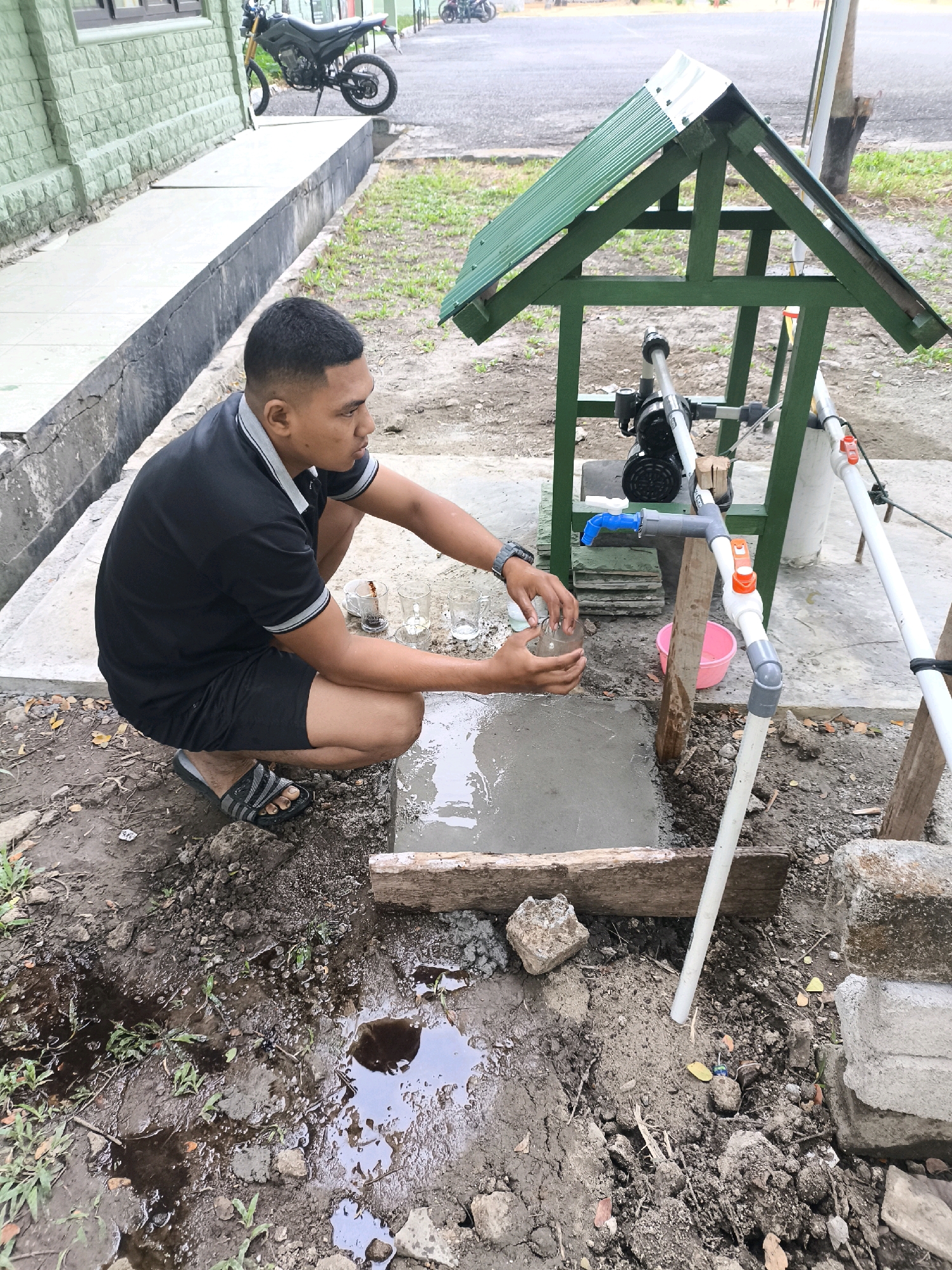 An outdoor scene depicts a young man squatting beside a small, makeshift water pump system. He is wearing a black polo shirt and black shorts, with black and gray Adidas sandals. The man is holding a clear glass in his right hand, appearing to be collecting water. The water pump system consists of a green wooden frame supporting a motor and pipes, with a blue faucet. A pink plastic bucket is positioned near the faucet. The ground is a mix of soil, cement, and some puddles of water. In the background, a motorcycle is parked near a building with a light green brick facade. The overall environment suggests an outdoor area in a rural or semi-urban setting, likely in Unknown, Indonesia.