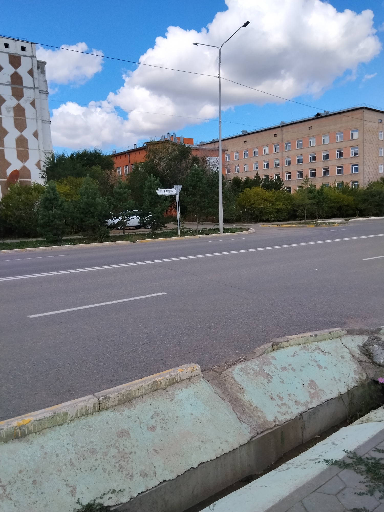 The image shows a street scene in Kokshetau, Kazakhstan, likely on a bright, cloudy day. In the foreground, a paved road with white lane markings is visible. To the right, a concrete drainage ditch runs alongside the road. In the midground, a white car is parked behind a line of trees. Beyond the trees, a multi-story brick building with numerous windows stands, appearing to be an apartment building or a public institution. To the left, a distinctive building with a geometric pattern of white and brown tiles is partially visible. A street lamp with two lights stands on the side of the road. A street sign with text in Cyrillic script is also present, indicating a location within the city. The sky is blue with scattered white clouds.