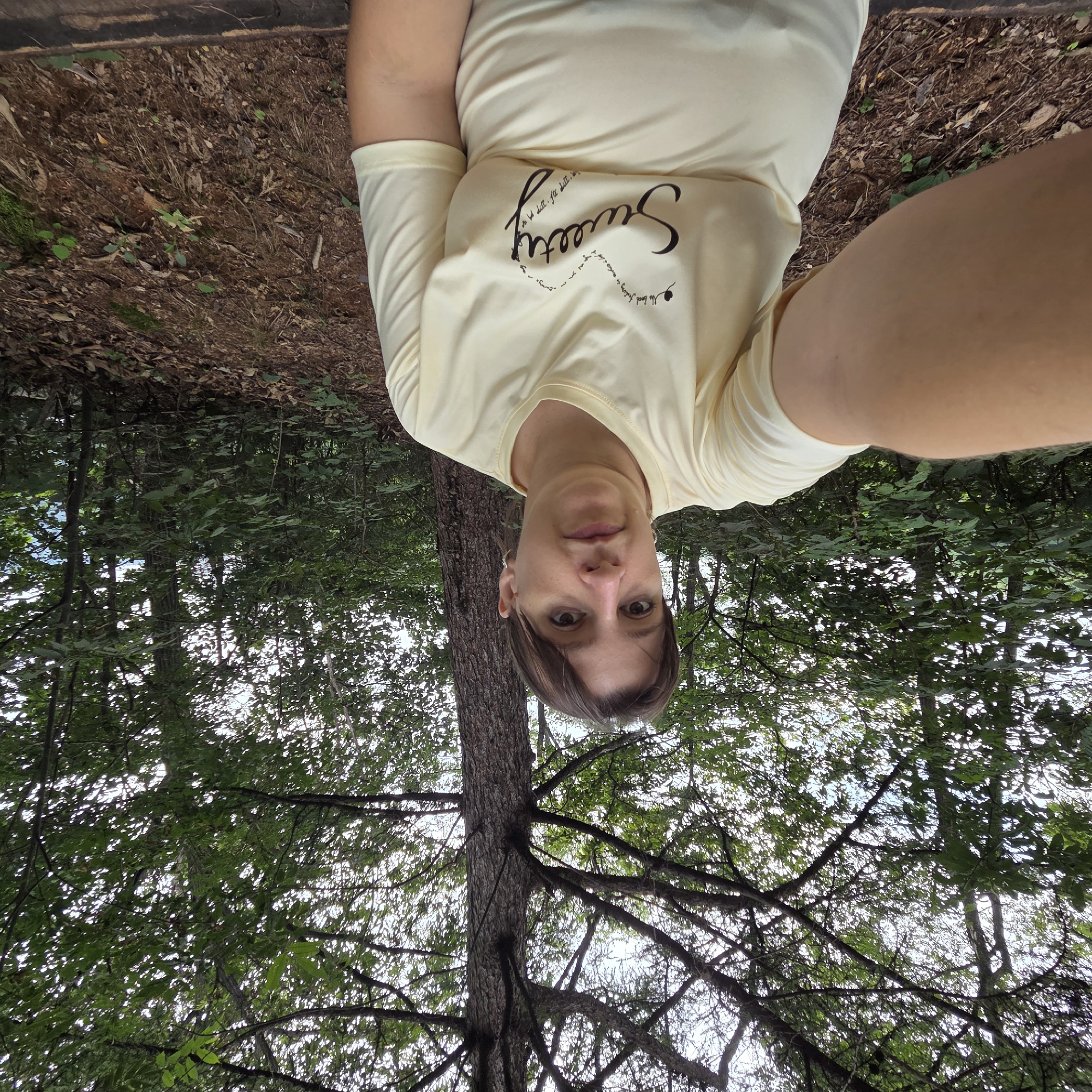 A selfie is taken by a person with short brown hair and fair skin, wearing a light yellow t-shirt with a graphic design that includes the word "Sweety" written in cursive. The person is seated outdoors in a wooded area, with a tree trunk visible behind them. The ground is covered with dry leaves and some greenery. The background consists of lush green trees and foliage, with the sky visible through the canopy. The overall impression is of a casual outdoor setting, possibly a park or a hiking trail. The location is identified as Gapcheon-myeon, South Korea.