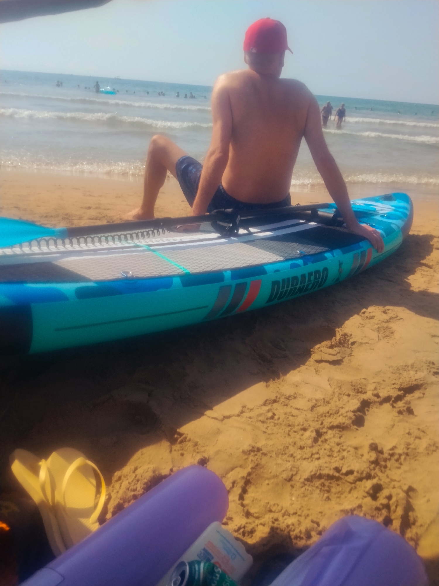The image depicts a scene on the beach in Orpesa, Spain. In the foreground, a turquoise and black paddleboard rests on the sand, with its side prominently displaying the text "DURAAERO". A person wearing a red baseball cap and dark blue swimming shorts is seated on the paddleboard, facing away from the camera, with their legs extended. The person's bare torso is visible. The background shows a sandy beach leading to the sea, with gentle waves breaking on the shore. Several other people are visible in the water, some appearing to be swimming or wading. The sky is clear and bright. In the immediate foreground, out of focus, are some items of beach gear including a yellow flip-flop and purple objects, possibly flotation devices, along with a can.