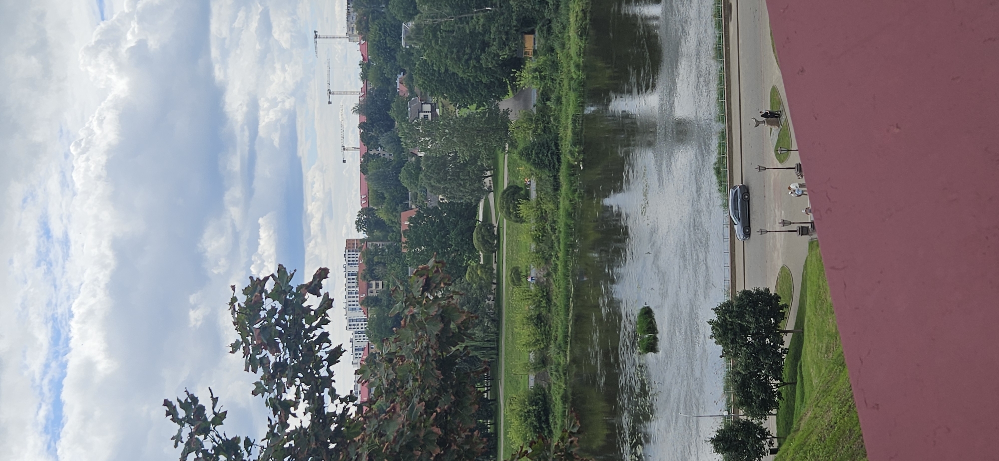This image, likely taken from an elevated viewpoint in Hrodna, Belarus, depicts a panoramic view of a city's landscape. In the foreground, a reddish-brown structure dominates the bottom portion of the frame. Beyond this, a gently sloping grassy area leads down to a paved pathway. A dark grey sedan is parked on the pathway, which runs alongside a body of water, likely a river given the context of Hrodna being situated on the Neman River. Several streetlights are visible along the pathway, and a small group of people, appearing to be two adults, are standing near a decorative monument or sculpture.
Across the water, a lush green embankment is covered with trees and shrubs, suggesting a park or recreational area. Further in the distance, a line of buildings with red roofs and modern apartment complexes with white facades are visible, interspersed with dense foliage. Three construction cranes are prominently displayed against the sky, indicating ongoing development in the city. The sky is filled with dramatic, partly cloudy formations. The overall scene conveys a sense of urban life integrated with natural elements, with a clear indication of both established infrastructure and new construction.