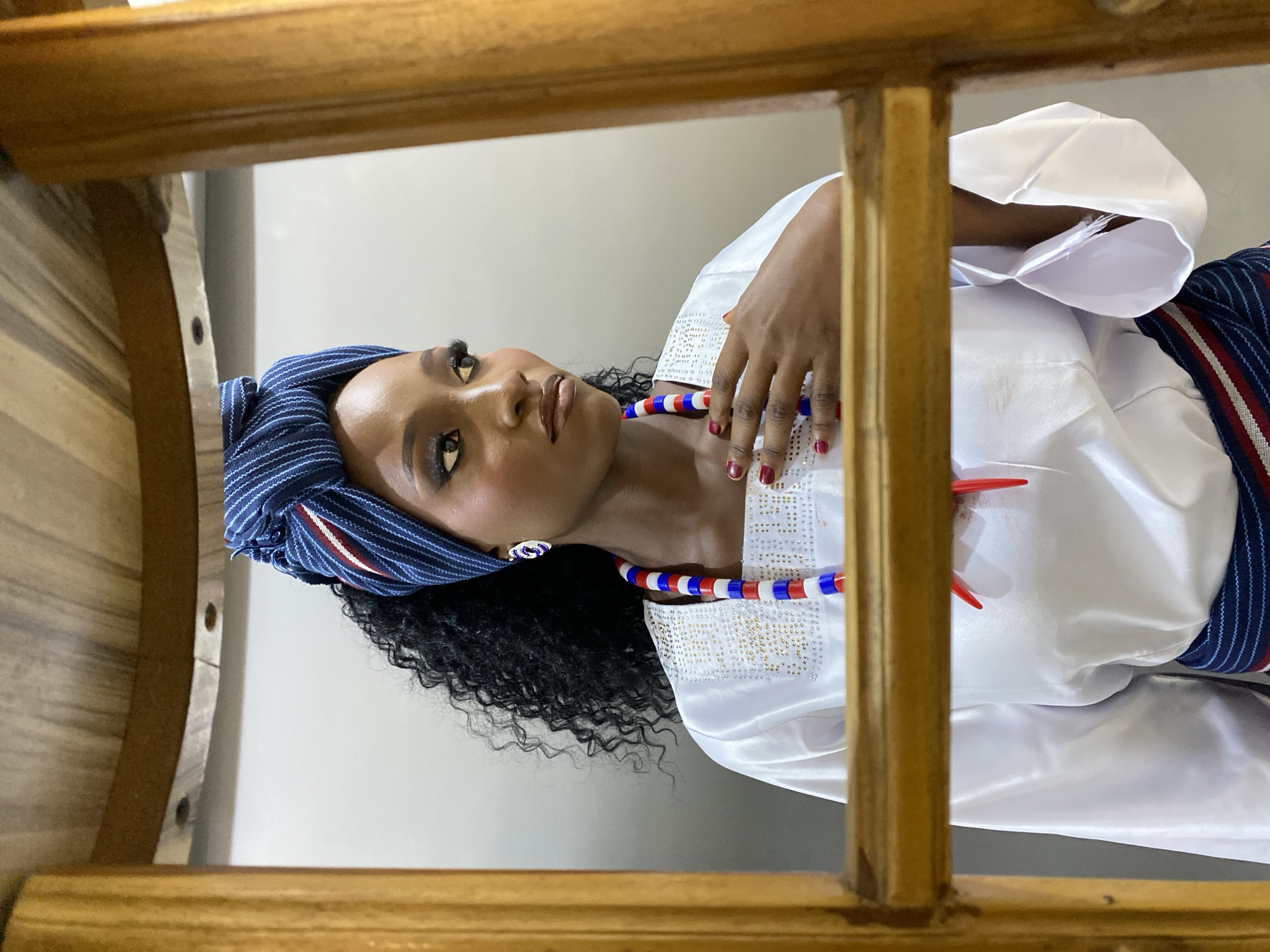 A young woman with dark, curly hair, styled in a blue and white striped headwrap, gazes to her right. She wears a white, long-sleeved garment adorned with gold embellishments around the neckline, and a multi-colored beaded necklace. Her left hand rests on her chest, with dark red nail polish visible. She is framed by a wooden structure, possibly part of a table or chair, and the background is a plain, light-colored wall. The image appears to be taken indoors. There is no information within the image to suggest it is located in Gura Top, Nigeria.