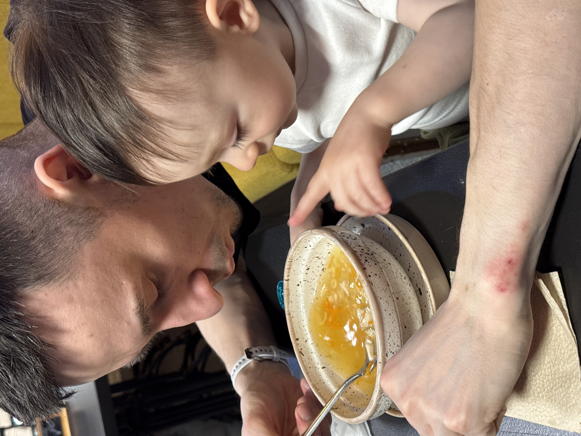 A close-up, indoor shot shows a fair-skinned adult male holding a bowl of soup for a young child. The man has short, dark hair and is wearing a light-colored digital watch on his left wrist. He is holding a bowl of broth-based soup with noodles. A metal spoon is inside the bowl. The child, with short brown hair and wearing a white shirt, is close to the bowl, with one finger pointing towards the soup. The man appears to be assisting the child with eating the soup. The man has an injury on his right wrist. The background is dark. The location cannot be determined from this image.