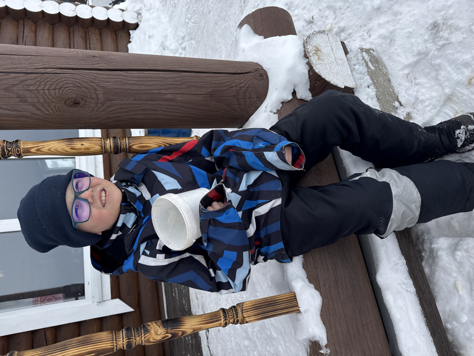 A child, appearing to be male, is seated outdoors on a snow-covered horizontal wooden log in Crokhovo, Russia. The child wears a dark blue knit beanie, eyeglasses with dark frames, a multi-patterned winter jacket in shades of blue, black, white, and red, and dark snow pants with lighter grey accents on the shins. Black winter boots are partially visible. The child holds a white, cylindrical plastic bucket in their left hand and gazes towards the upper right with a slight smile. Behind the child is a log cabin-style building constructed from dark brown logs, with a white-framed window partially visible on the left. Two prominent vertical wooden posts frame the child: a carved, lighter-colored post to the left and a thicker, darker post to the right, both appearing to be structural elements of a porch or entrance. The ground and horizontal surfaces are covered in fresh snow.