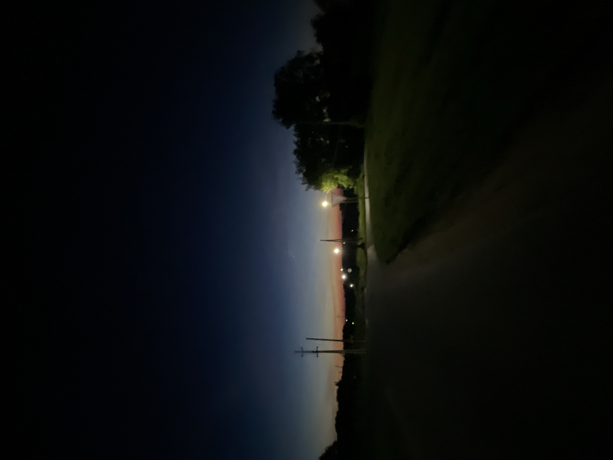 The image depicts a dark, rural road at dusk. The sky transitions from a deep indigo at the top to a faint orange and pink hue near the horizon. Several utility poles with wires are visible along the left side of the road, and a few streetlights cast pools of light on the right. Trees and brush line the sides of the road, creating silhouettes against the darkening sky. The road itself is paved and appears to be in good condition. The overall impression is one of tranquility and solitude, characteristic of a rural setting. The location is specified as Krasnye Baki, Russia.