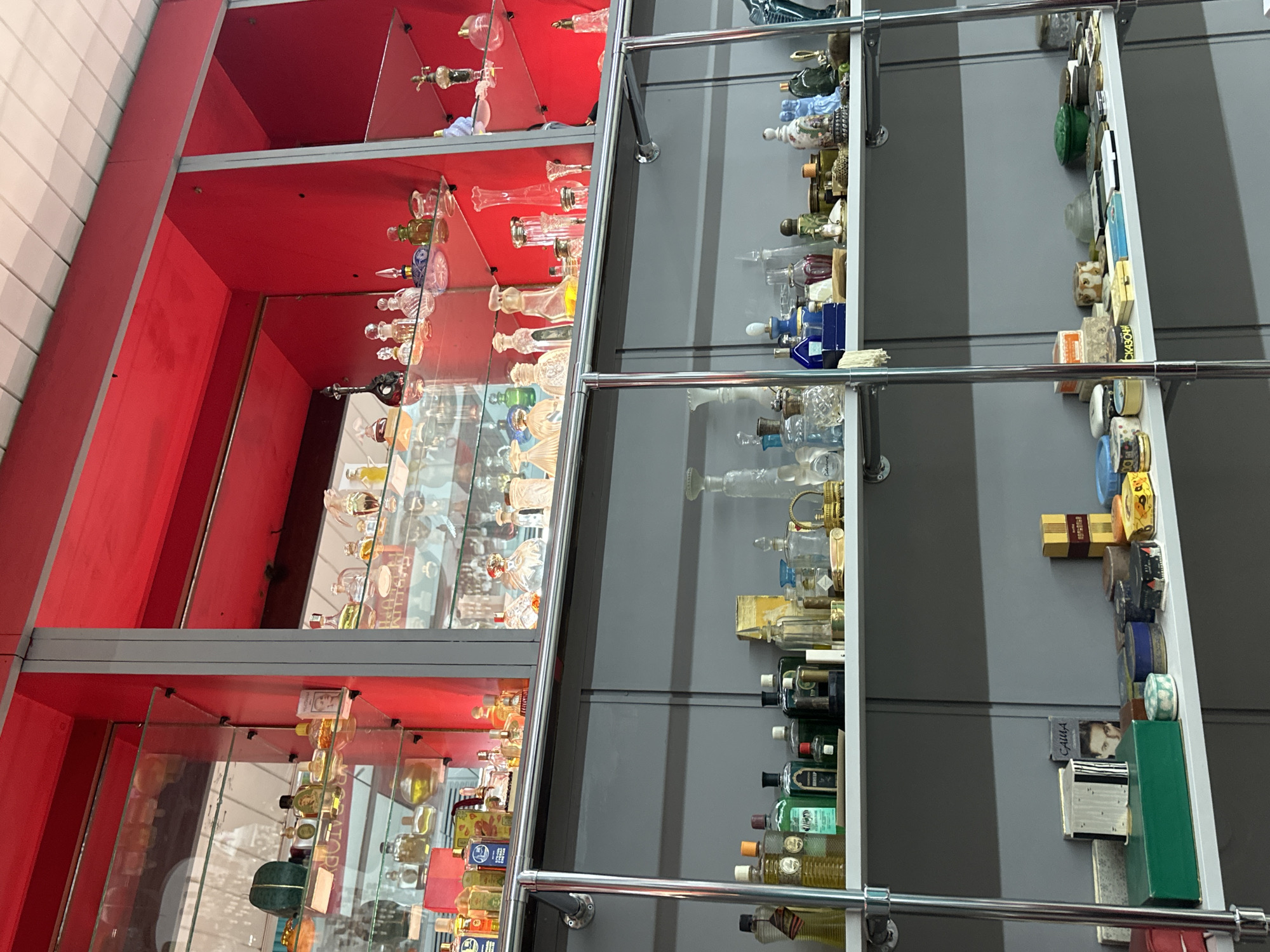 The image shows a brightly lit display of vintage perfumes and cosmetics on shelves in what appears to be a retail store or museum. The shelves are made of glass and metal, and they are illuminated by overhead lighting. The background is a bright red, suggesting a decorative or thematic element of the display. The items on display are arranged in an orderly fashion, with a variety of bottles, boxes, and containers. The focus is on the colorful and intricate designs of the perfume bottles, many of which appear to be made of glass. The overall impression is one of a curated collection, showcasing a history of perfumery and beauty products. There are no people or animals visible in the image.