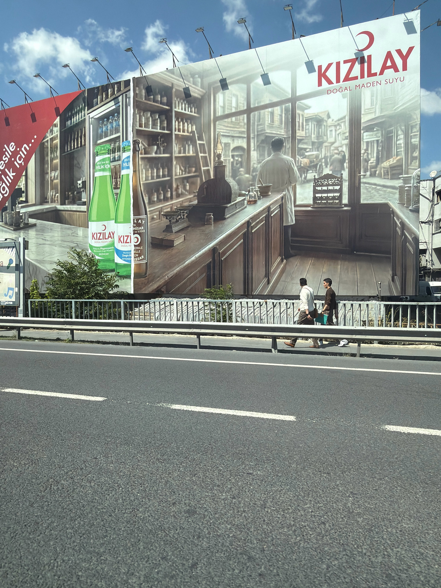 An outdoor, wide shot captures a large billboard advertising Kızılay Doğal Maden Suyu, situated above a highway in Zeytinburnu, Turkey. The billboard displays a historical scene with shelves of bottles, a counter, and a person in a white coat looking out a window onto a street with horse-drawn carriages. Two men walk along a sidewalk below the billboard, separated by a metal railing and the highway. The sky is blue with scattered clouds, and the road is asphalt with white lane markings.