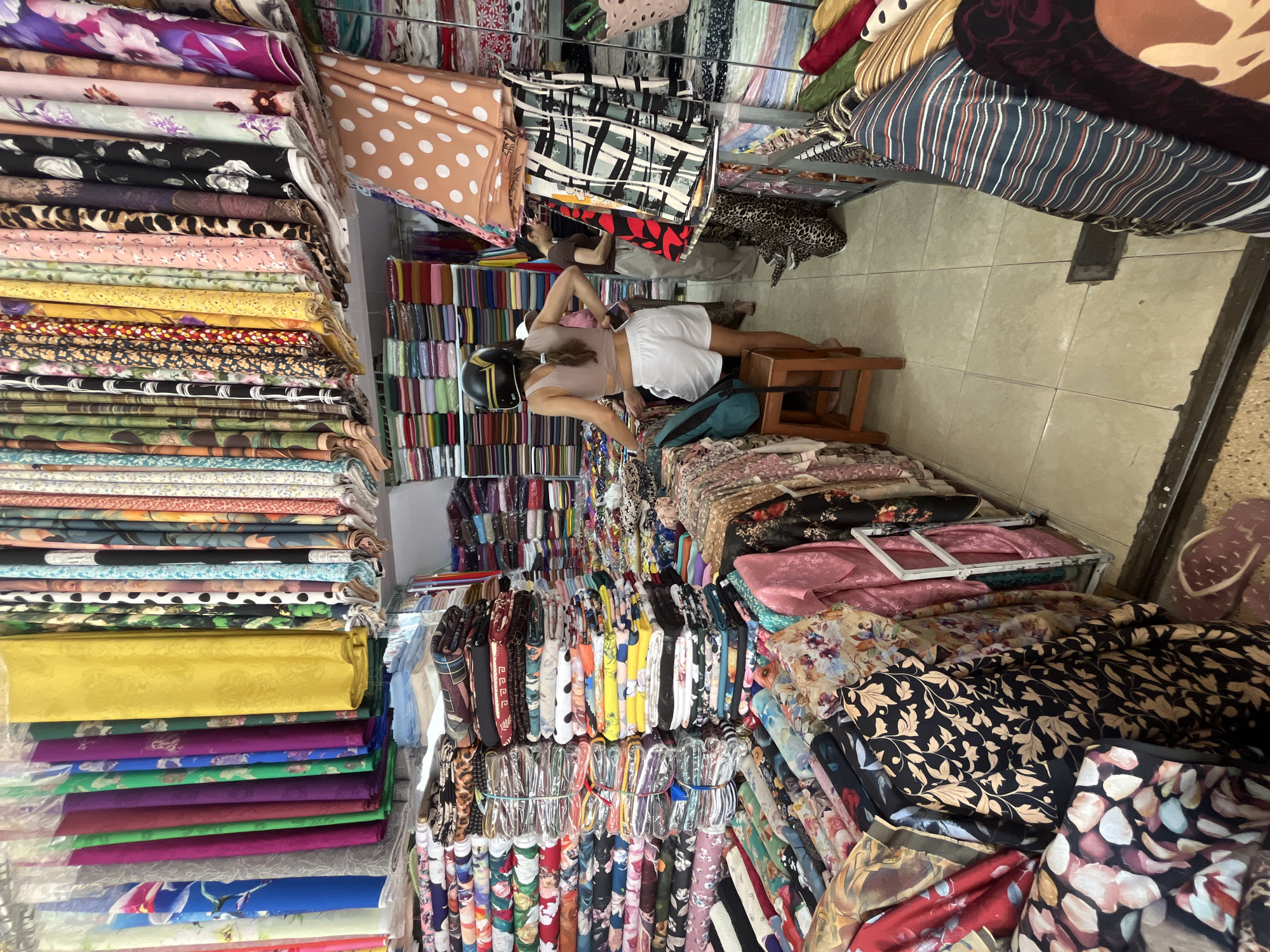 A photograph taken indoors shows a fabric shop in Nha Trang Ward, Vietnam. The shop is densely packed with bolts and rolls of fabric stacked and hung along the walls. The fabrics vary in color and pattern. In the center of the shop, a woman wearing a helmet lies on a stool. In the background, another person is lying on fabric. The floor is tiled.