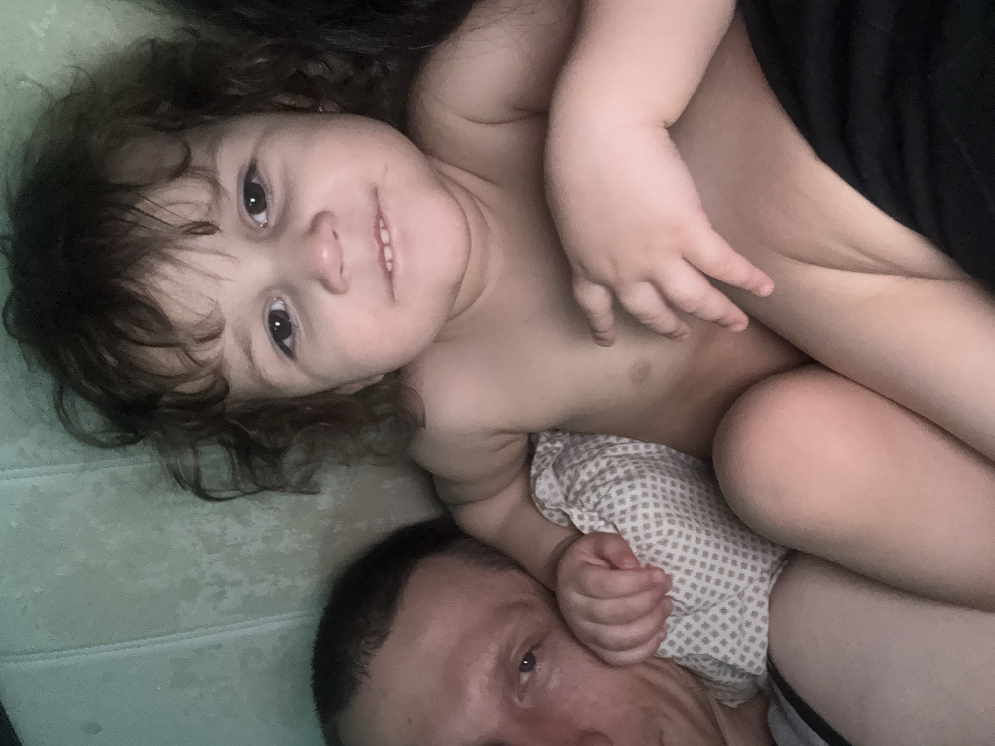 The image displays a tender, intimate moment between an adult and a young child, captured from an overhead perspective, likely a selfie. A young child, appearing to be a toddler or preschooler, lies on their back, positioned on top of or beside an adult. The child has dark, curly hair and large brown eyes, and is looking directly at the camera with a bright, happy smile, revealing small teeth. The child is shirtless, showing bare arms and a part of their torso.
Beneath the child, and partially visible in the lower section of the frame, is an adult. Only the top of the adult's head, face, and a shoulder are seen. The adult has short dark hair and is also looking towards the camera with a gentle, soft expression, appearing to be content. The child's left arm and hand are resting on a patterned fabric, which also extends beneath the child's lower body and near the adult's head. This fabric is light-colored with small, dark, square-like patterns, possibly a pillow or blanket.
The background consists of a light green or blue textured surface, likely a mattress or bedsheet, under the child's upper body and head. A dark, solid-colored fabric (possibly a blanket or clothing) is visible behind the child's back. Another striped garment, grey and light-colored, can be seen near the adult's leg.
The overall scene conveys a feeling of affection, comfort, and happiness between the adult and child. The lighting suggests an indoor setting, but the specific time of day is not discernible. There are no visible location cues within the image itself to confirm the stated location of Tobolsk, Russia, nor is any text visible.