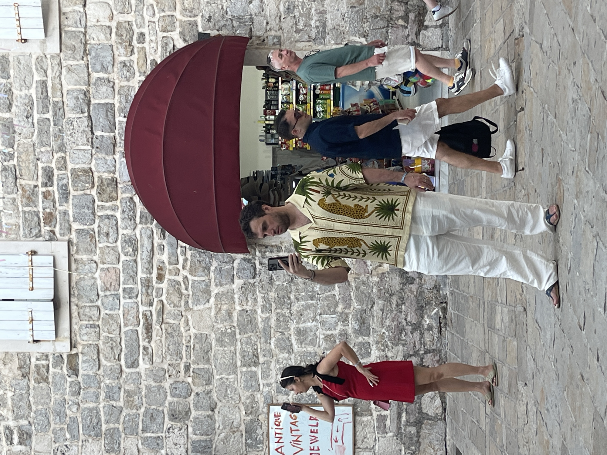 In Budva, Montenegro, in front of a stone-walled building with a red awning and a window with light blue shutters, three men and one woman are captured in a candid shot. The woman, on the left, wears a red dress and holds a smartphone. To her right, a man in a light-colored shirt with a leopard print and white pants is also holding a smartphone. Further right, a man in a dark blue shirt and white shorts walks with a backpack, followed by another man in a green t-shirt and white shorts. The ground is paved with cobblestones.