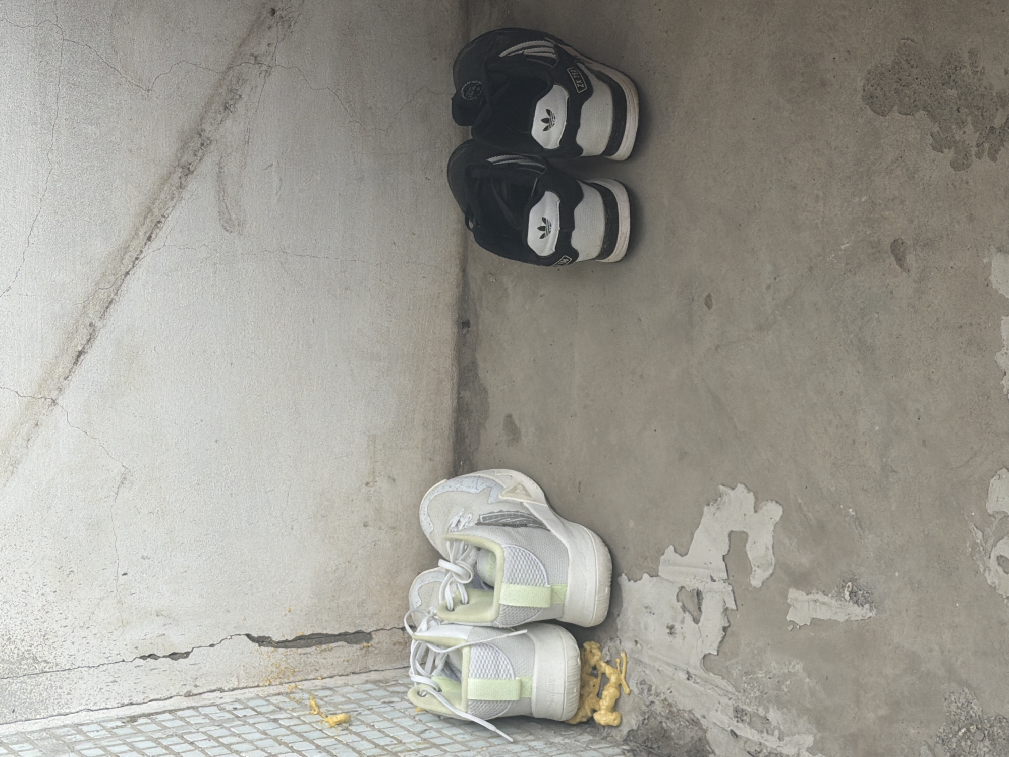 Location: Likely an exterior corner space in Chaikovsky, Russia, based on the urban architectural style and concrete construction.
Environment: A concrete structure with noticeable wear and tear. The corner has both smooth concrete and areas where the surface has peeled away, revealing the under-layer. A tile floor meets the base of the wall.
Objects:
* Two pairs of sneakers, stacked. One pair of black and white Adidas sneakers is placed higher up. A pair of white sneakers is placed lower, closer to the base.
* Yellow foam debris is scattered near the base of the white sneakers and on the tile floor.
Action: Possible storage or abandonment of the sneakers in the corner. No people are visible.
Interactions: No interactions are visible.