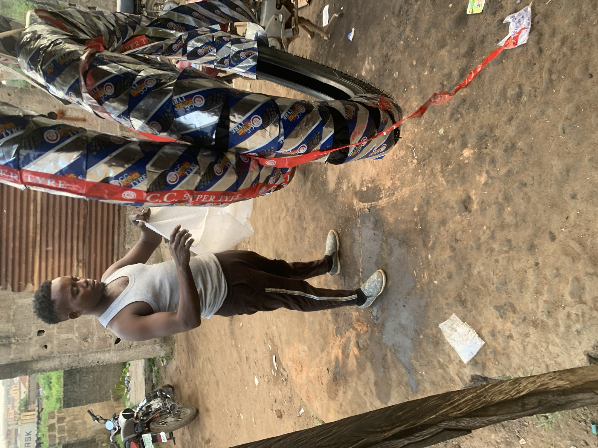 A young man is standing on a dirt ground, holding a piece of white plastic. He is wearing a white tank top and dark brown pants with a white stripe down the side, and grey and black shoes. He has short, dark hair and is looking down at the plastic. Behind him are several motorcycle tires wrapped in silver and blue packaging with red trim, secured with red plastic ties. The tires are stacked on top of each other and leaning against something out of frame. To the left of the man, a corrugated metal wall is visible, and behind that, some greenery and buildings suggest an outdoor or semi-covered setting. To the man's right, a small motorcycle is parked on the ground. The overall scene appears to be a workshop or a storage area in an outdoor environment. Visible text on the tire packaging includes "C.C. SUPER TYRE". The lighting suggests it is daytime.