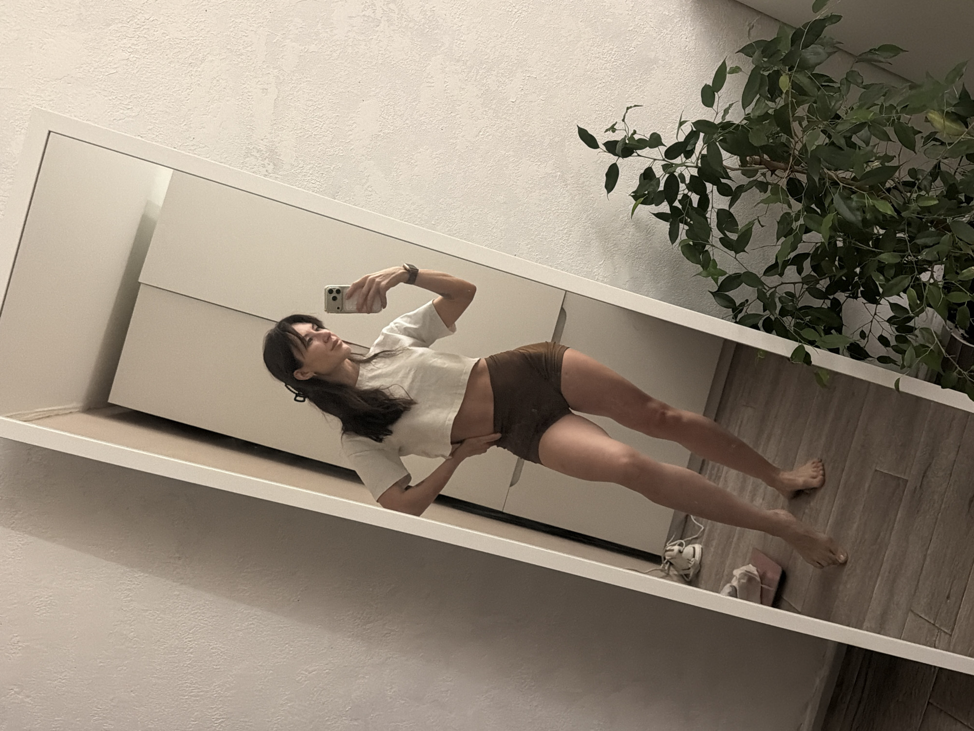 A woman is taking a selfie in a mirror. She is wearing a white crop top and brown shorts. The mirror reflects a wardrobe, a plant, shoes, and a wooden floor. The background includes a white textured wall. The location is likely indoors. There is no indication the image was taken in Perm, Russia.