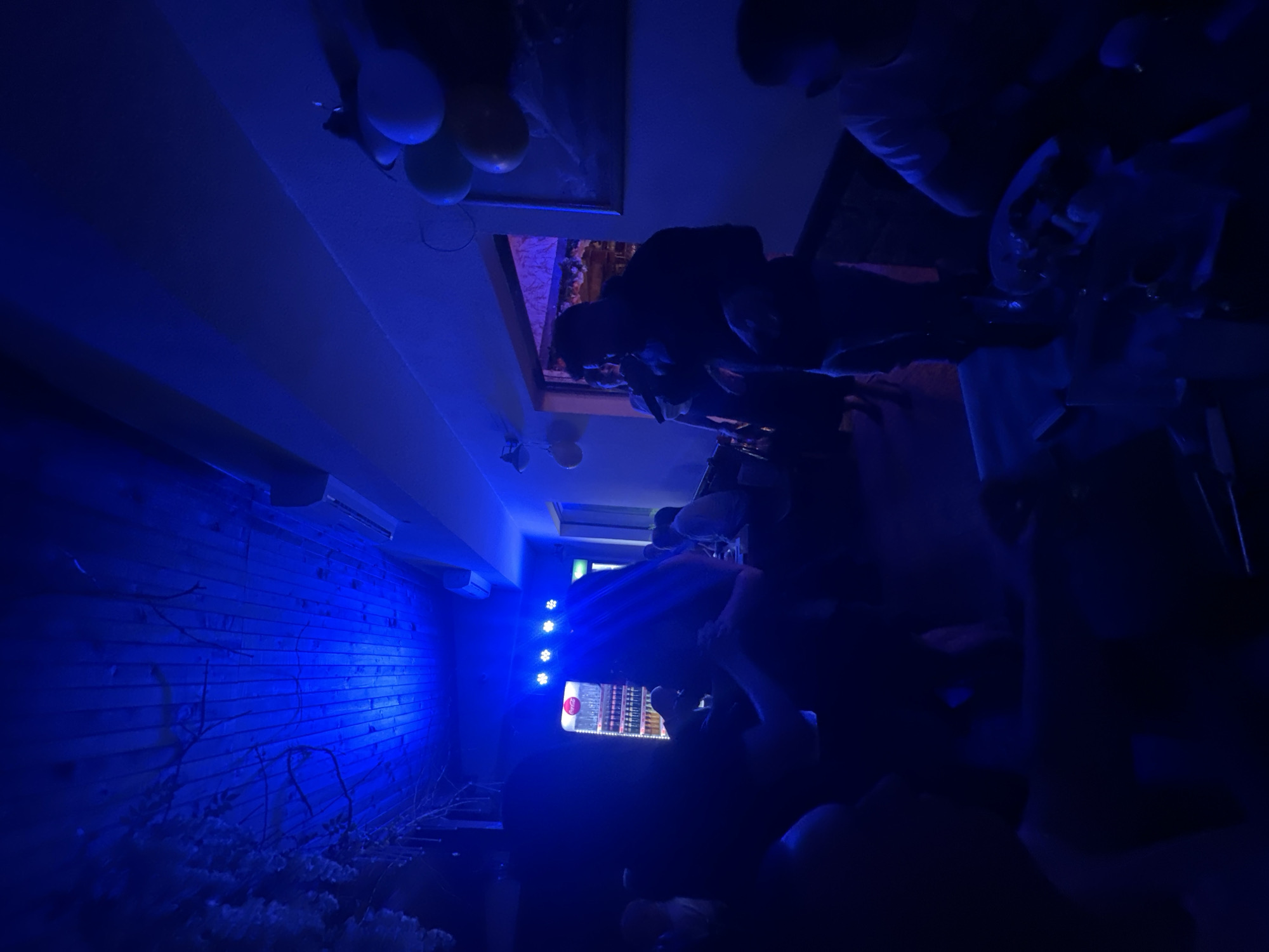The image shows an indoor scene with a dark ambiance illuminated by blue lighting. A brick wall is visible on the left. In the background, there are a few people gathered in what appears to be a room. There are balloons hanging from the ceiling. Based on the location, the image is likely taken in Panagyurski kolonii, Bulgaria.