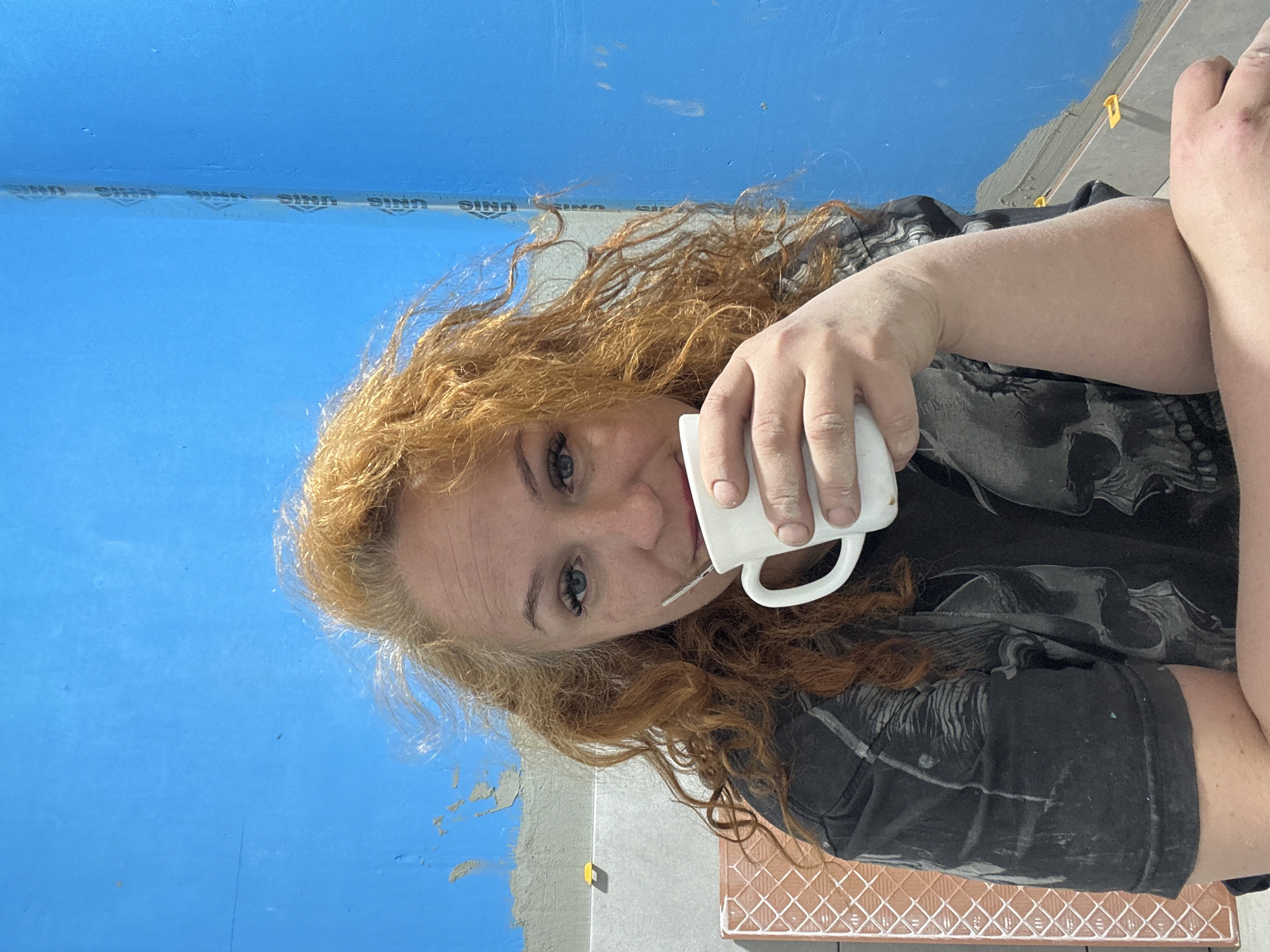 A person with curly red hair is holding a white mug to their lips. They are wearing a dark grey t-shirt with a skull graphic. The background includes a blue wall partially covered with grey material, potentially in the process of tiling. A corner reveals tiled flooring. The location appears to be a domestic interior, potentially under renovation. Specific city or geographic details beyond a generic interior space are not discernible from the image.
