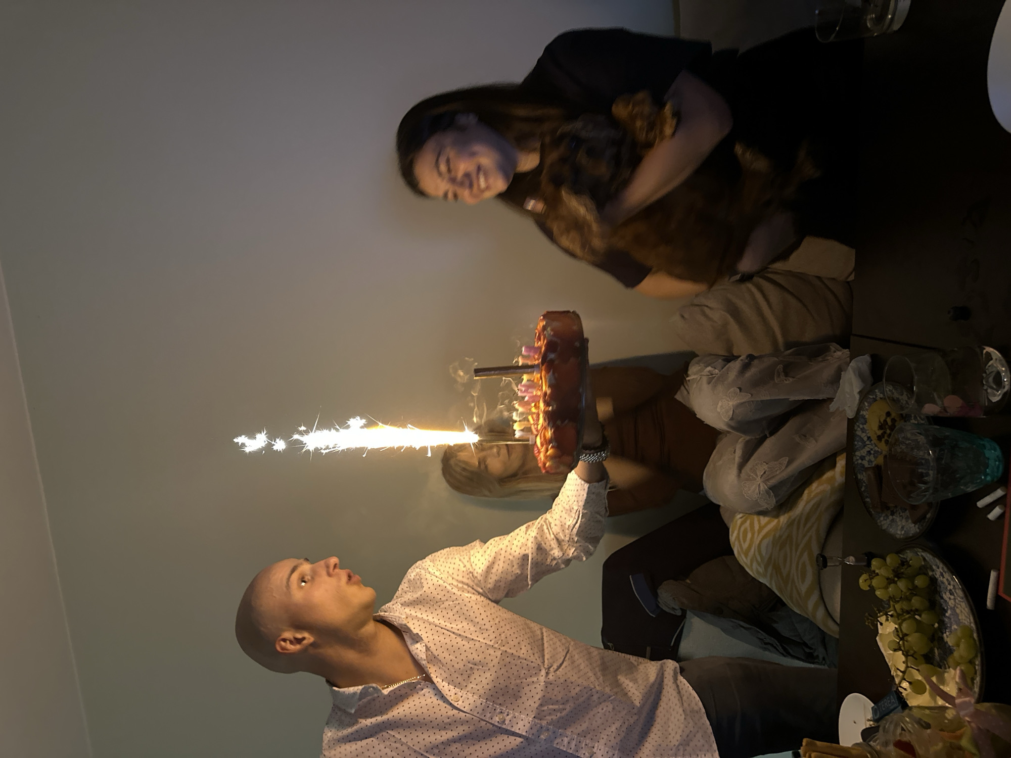 This image captures an indoor celebration, likely a birthday party, taking place in the evening or at night in Sofia, Bulgaria. The scene is shot from a low angle and is oriented vertically (originally rotated).
In the foreground, a young man with a shaved head, wearing a light-colored, polka-dotted long-sleeved shirt, holds a multi-layered cake. A large, brightly burning sparkler is inserted into the cake, illuminating the immediate area with its shower of sparks. The man looks up with an engaged expression towards the sparkler.
Behind the cake and to the right, a woman with long dark hair, wearing a dark top, smiles as she holds a small, brown, furry dog. She also appears to be observing the sparkler. Another person with lighter hair and a brown top is partially visible behind the cake, suggesting more people are present at the gathering.
The setting appears to be a cozy indoor space, possibly a living room or dining area. A dark table to the right of the cake is laden with party items, including plates, empty glasses (one with a blue tint), a plate of green grapes, and various other snacks or wrapped items. Faint text "TERRA" is visible on a small white object on the table. The background features a plain light-colored wall and hints of soft furnishings. The overall mood is festive and joyful, centered around the illuminated cake.