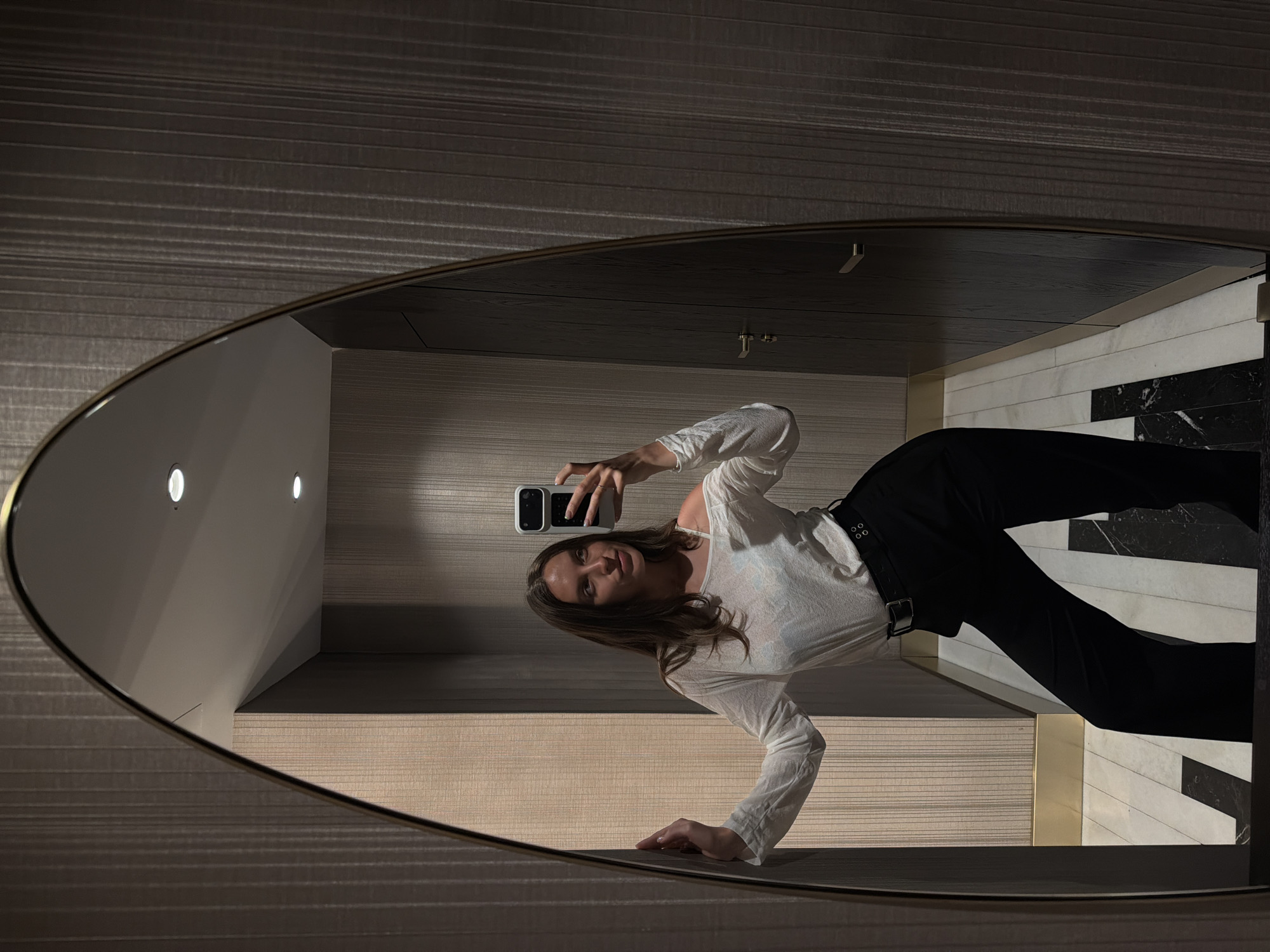 A young woman is depicted taking a mirror selfie in an indoor setting, likely a hallway or room, in Budva, Montenegro. She stands with her left hand resting against a textured wall and holds a white smartphone with a prominent black camera module in her right hand to capture her reflection. She has long, brown hair and is wearing a loose-fitting, off-white long-sleeved top, revealing a light blue bra strap on her right shoulder, paired with black high-waisted trousers and a black belt with a silver buckle and two silver grommets.
The mirror, framed in a thin gold border, has a unique, curved top edge and reflects a sophisticated environment. The walls within the reflection are covered in textured wallpaper with vertical ridging; a lighter beige section is visible on the left, transitioning to a darker greyish-brown on the right. The ceiling is light-colored and features two illuminated recessed spotlights. The floor showcases a striking geometric pattern of black and white marble tiles. Gold-colored metallic baseboards run along the bottom of the walls. On the right side of the reflection, a dark wooden door or cabinet with minimalist horizontal gold handles is visible.