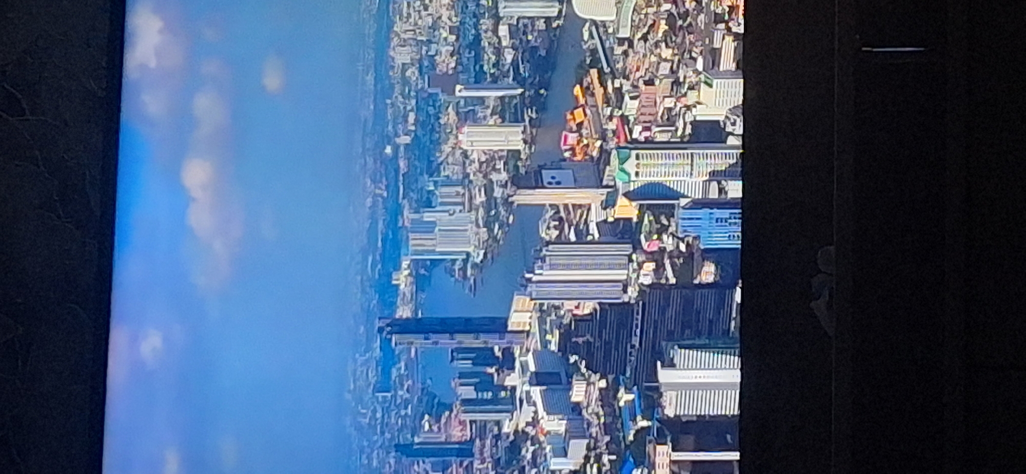 This is a high-angle aerial shot of a dense urban landscape with numerous high-rise buildings. A wide river flows through the center of the city, with several bridges visible. The sky is a clear blue with scattered white clouds. The image is slightly blurry and pixelated, suggesting it was captured from a distance or on a lower-resolution screen. There are no discernible individuals or specific landmarks that would identify the city. The lower portion of the image shows a dark foreground that appears to be the bottom of a screen or display. There is no evidence to suggest this image depicts the city of Bila Tserkva, Ukraine.