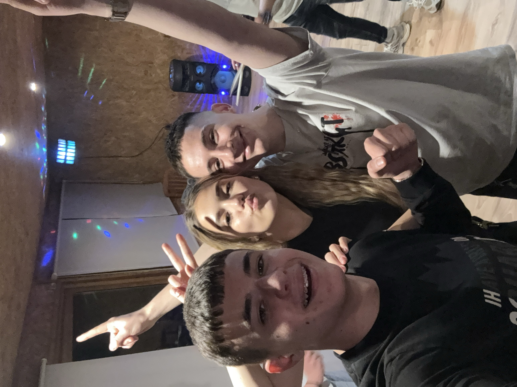 Three people are posing for a selfie indoors. A young man with dark hair and braces is in the foreground, smiling and making a peace sign with his left hand, while extending his right index finger towards the camera. A young woman with light brown hair is behind him, puckering her lips for a kiss. A second young man is above the woman, wearing a light grey t-shirt and holding up his right fist, also smiling. The background includes a wall with wood paneling, disco lights, and a speaker. The environment suggests a casual, possibly party setting. The location is Strashimirovo, Bulgaria.
