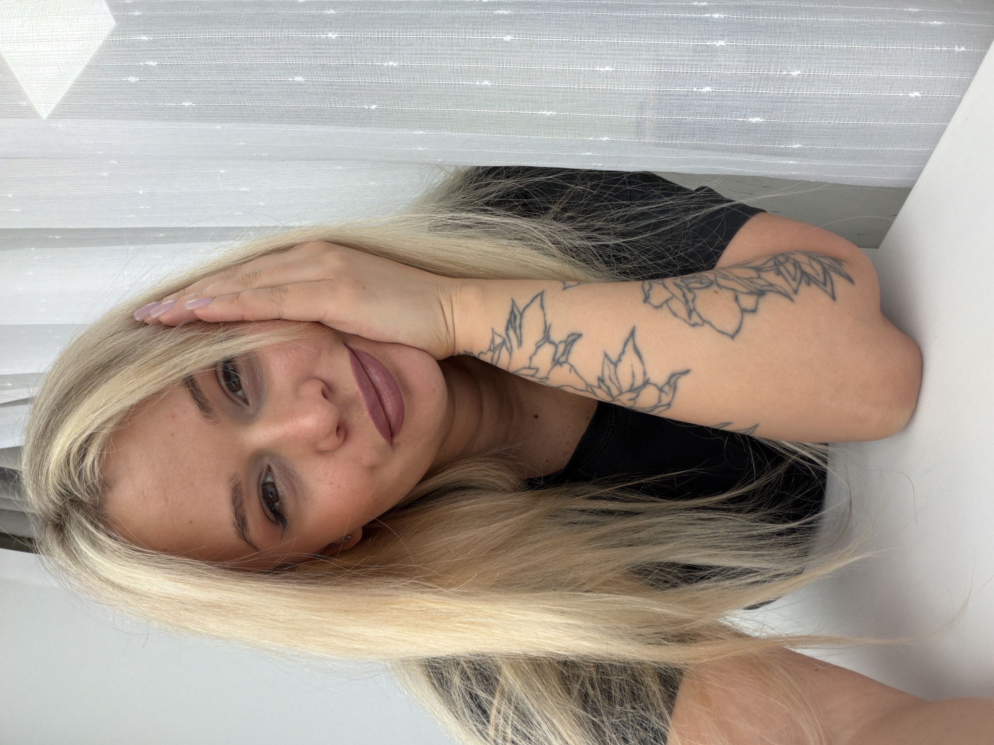 A portrait of a young woman with long, straight blonde hair is captured with a significant tilt, making her head appear rested towards the left side of the frame. She has light-colored eyes and looks directly at the camera with a subtle, relaxed smile. Her skin is light-toned, and she wears eye makeup and matte, reddish-brown lipstick, complemented by a small piercing below her lower lip. She is dressed in a black top. Her left arm, positioned in the foreground, is bent at the elbow, with her hand gently resting on the side of her head and hair. This arm features several detailed outline tattoos of botanical elements, such as flowers and leaves. Her fingernails are painted with a light lavender or pink polish.
The background consists of a textured white sheer curtain or blind on the left, displaying horizontal stripes and small, regularly spaced white dots. To the right, a plain white surface, possibly a wall or table, completes the minimalist setting. The lighting is soft and diffused, creating an evenly lit scene without harsh shadows, indicative of an indoor environment. There is no visible text within the image. The overall mood conveyed is calm and engaging. This media file originates from Sarapul, Russia.