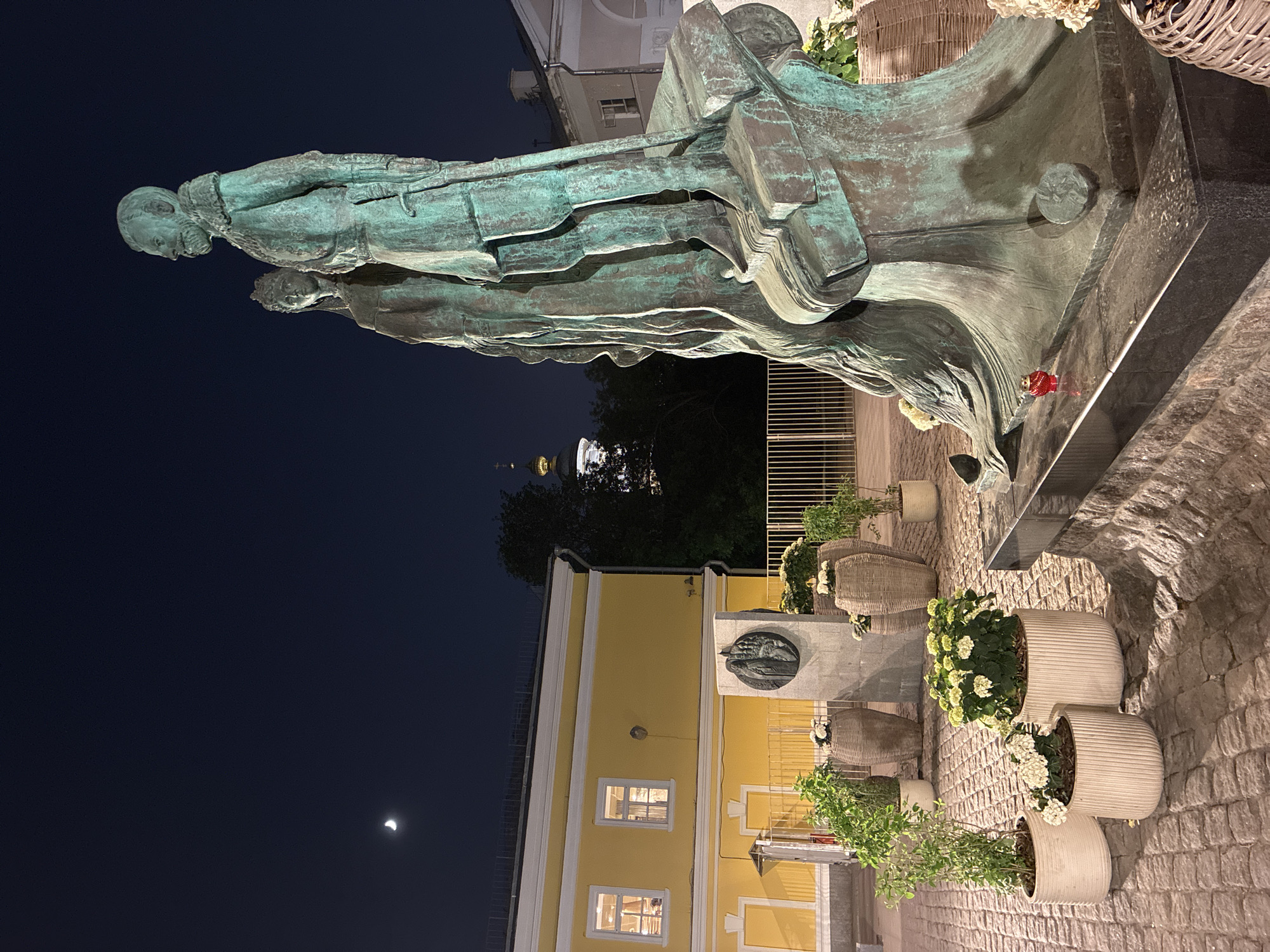 The image depicts a nighttime scene featuring a bronze statue of a man holding a book, positioned in front of a yellow building. To the left is a dark, partially visible night sky with a faint moon. The statue rests on a dark stone base adorned with floral arrangements in pots. The yellow building behind the statue features illuminated windows. In the background, a church dome is faintly visible amidst the trees. The scene is set on a cobblestone surface. The statue is the monument to the Belarusian Educator, Philosopher and humanist Francysk Skaryna located in Moscow, Russia.