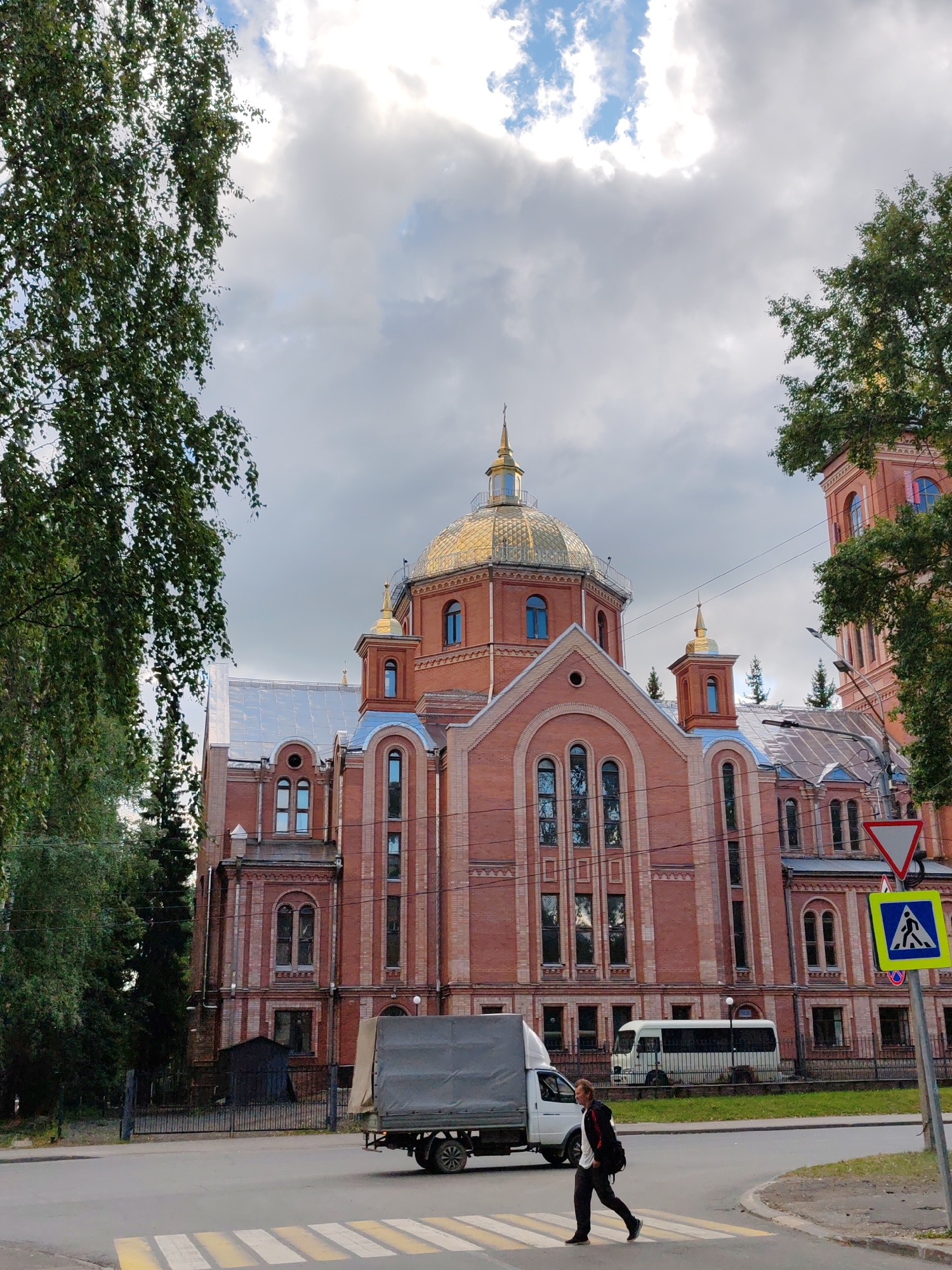 In an urban environment in Syktyvkar, Russia, a man is crossing a pedestrian street. The man is wearing dark pants and a white shirt. He has a backpack on his back. He is walking from right to left. There is a large, red brick building behind him. The building has a gold dome and several windows. The sky is cloudy. There is a truck in front of the building. A white bus is behind the truck. There are trees on the left side of the image. There is a pedestrian crossing sign on the right side of the image. The road has a yellow and white striped pedestrian crossing.