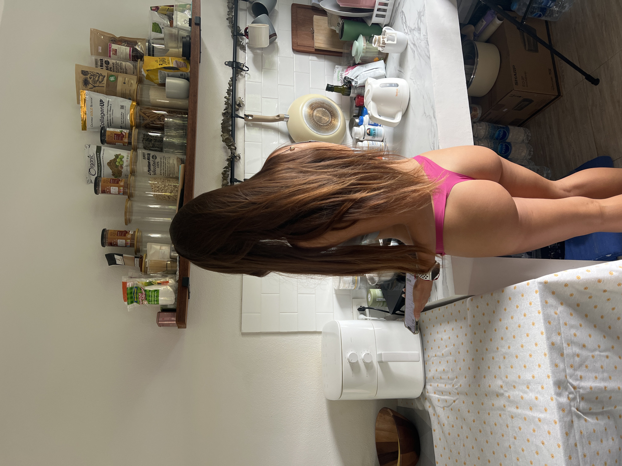 A young woman with long, brown hair stands in a kitchen, facing away from the camera. She is wearing a pink thong. The kitchen has white subway tile backsplashes, white countertops, and wooden shelves stocked with various food items and containers. To the left of the woman is a white air fryer and a wooden bowl. On the counter to her right are a white electric kettle, a mug with a coffee drip filter, and other items. The floor is a dark wood laminate. The room appears to be well-lit by natural light.