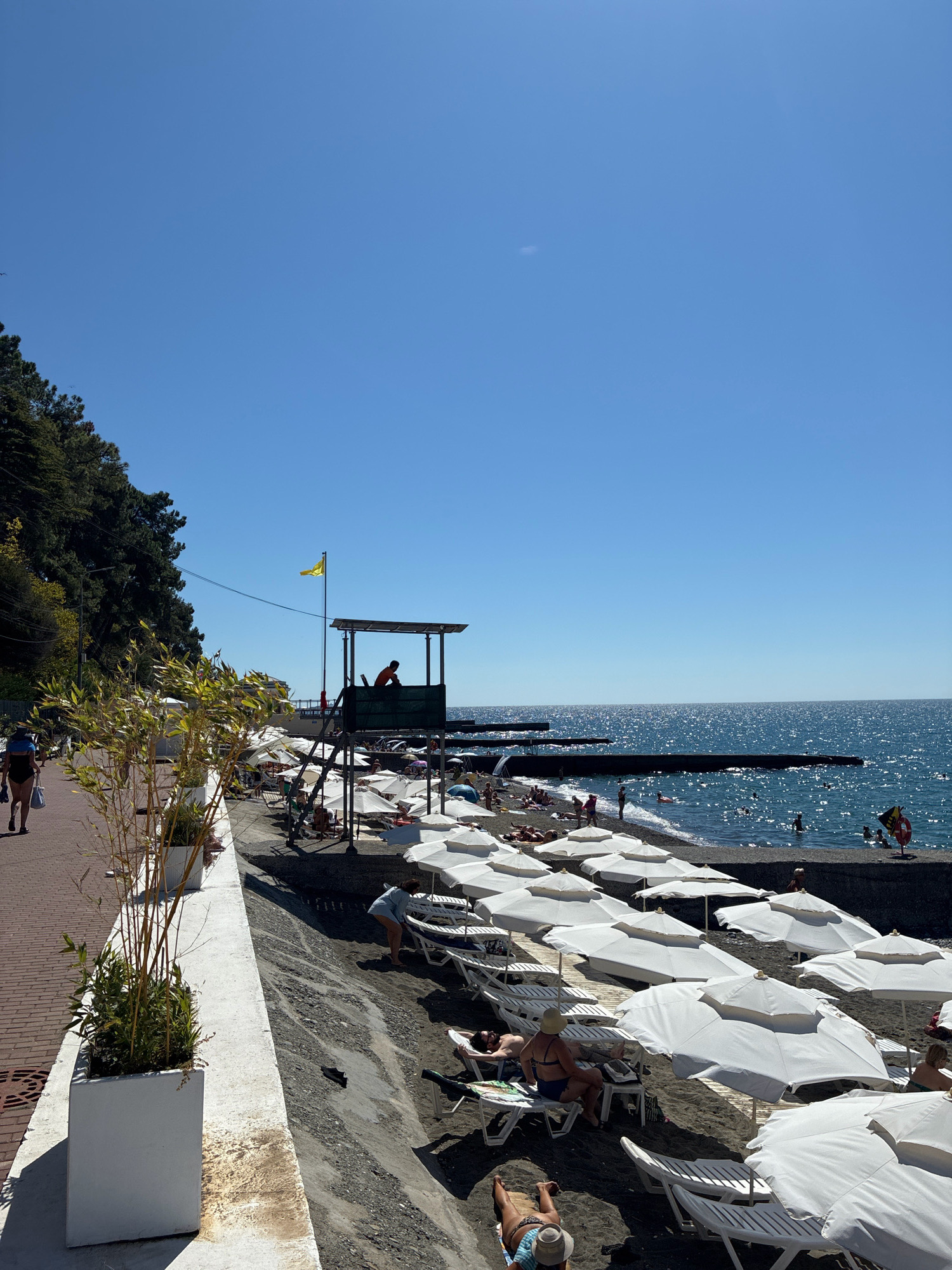 The image depicts a sunny day at a beach in Sochi, Russia. A paved walkway runs along the left side of the frame, lined with white planters containing tall, slender plants. A woman in a black swimsuit walks on the walkway, carrying a blue bag. Beyond the walkway is a pebble beach covered with numerous white lounge chairs and umbrellas. In the background, people are sunbathing and swimming in the clear blue sea. A lifeguard tower stands prominently on the beach, with a lifeguard visible inside. Several dark, rectangular structures extend into the water, possibly piers or breakwaters. The sky is a clear, bright blue. The overall scene is one of a popular tourist destination on a warm summer day.