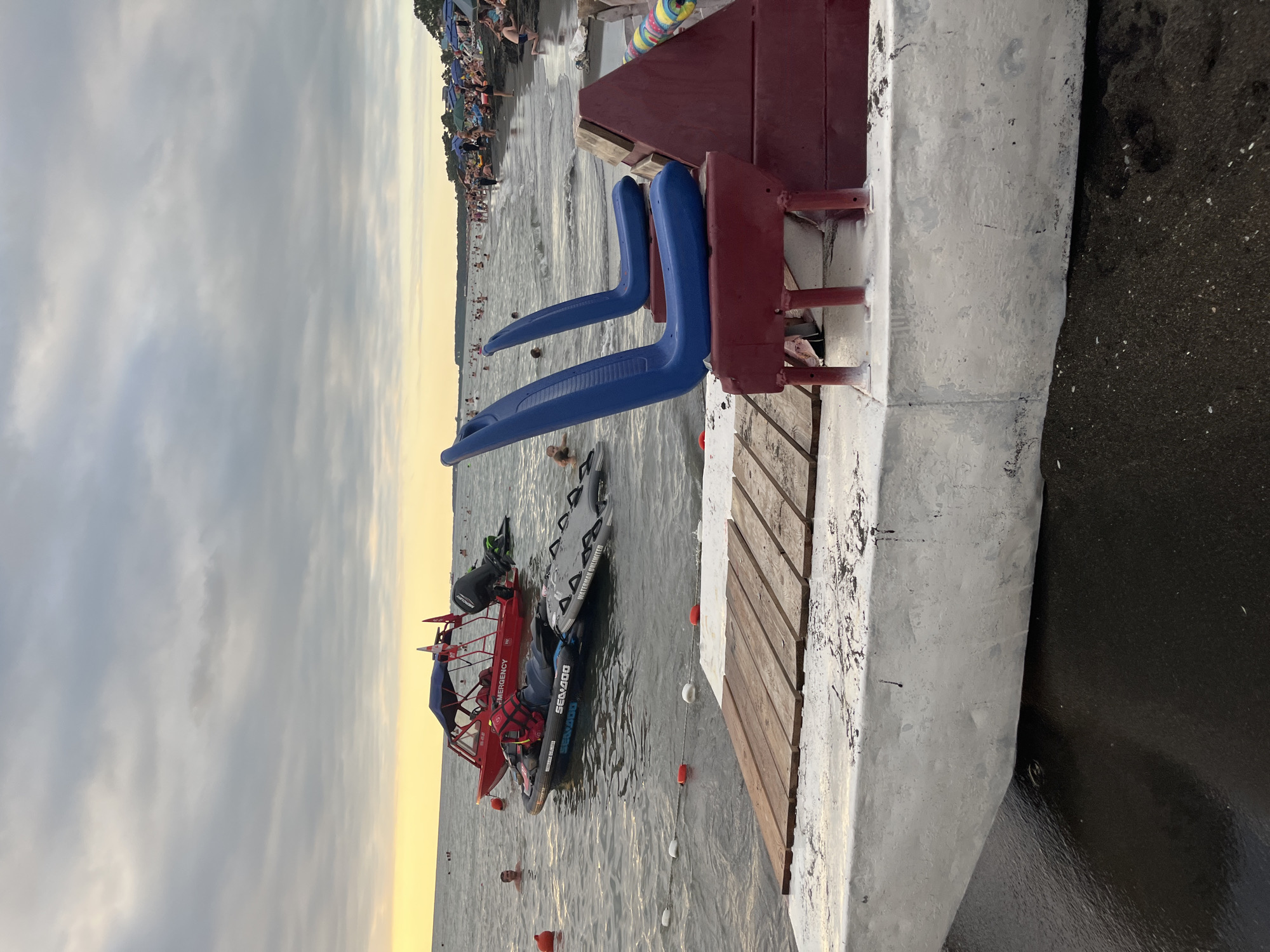 An outdoor, eye-level shot captures a portion of the beach in Ureki, Georgia, with a focus on a partially submerged pontoon boat in the foreground. The boat's hull, painted white with visible wear and tear, rests on dark, wet sand at the water's edge. A wooden deck is installed on the front of the pontoon, and two blue plastic seats are mounted on a red metal frame attached to the boat's rear. A red structure, possibly part of the boat's amenities or a docking station, is visible behind the seats.
In the background, the Black Sea is visible with moderate waves. Several buoys, red and white, are scattered in the water. Two boats are also present: a red emergency boat with a canopy and an outboard motor, and a black jet ski with a person in a red life vest lying on it. The water is populated with swimmers, and the shore is lined with beach umbrellas and sunbathers. The sky is overcast with dramatic clouds and a soft glow of the setting sun visible on the horizon.