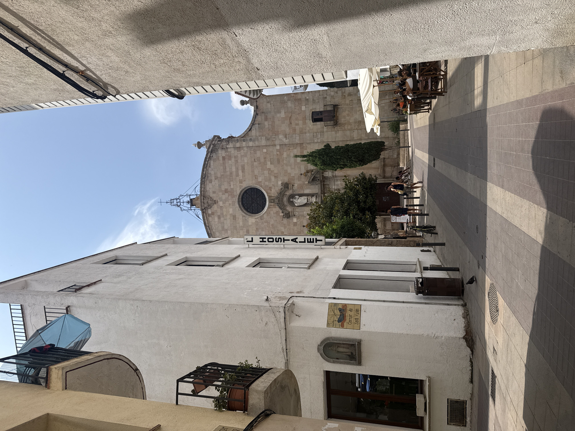 The image depicts a sun-drenched street scene in Tossa De Mar, Spain, likely within the historic Vila Vella. On the left, a white building features a balcony with a blue umbrella hanging from it. Below the balcony is a window and a small plaque with an image of the Virgin Mary and text that reads "Carrer de Sant Elm". A vertical sign on the building states "L'HOSTALET".
The street, paved with alternating light and dark grey square tiles, leads towards a prominent stone church with a circular window above its entrance and a decorative spire. Trees are visible on either side of the church entrance.
On the right side of the street, an outdoor cafe is set up with several tables and chairs under white umbrellas. Patrons are seated at the tables, enjoying drinks and conversation. People are walking along the sidewalk in the middle distance. The overall atmosphere is one of a bright, lively Mediterranean town on a clear day.