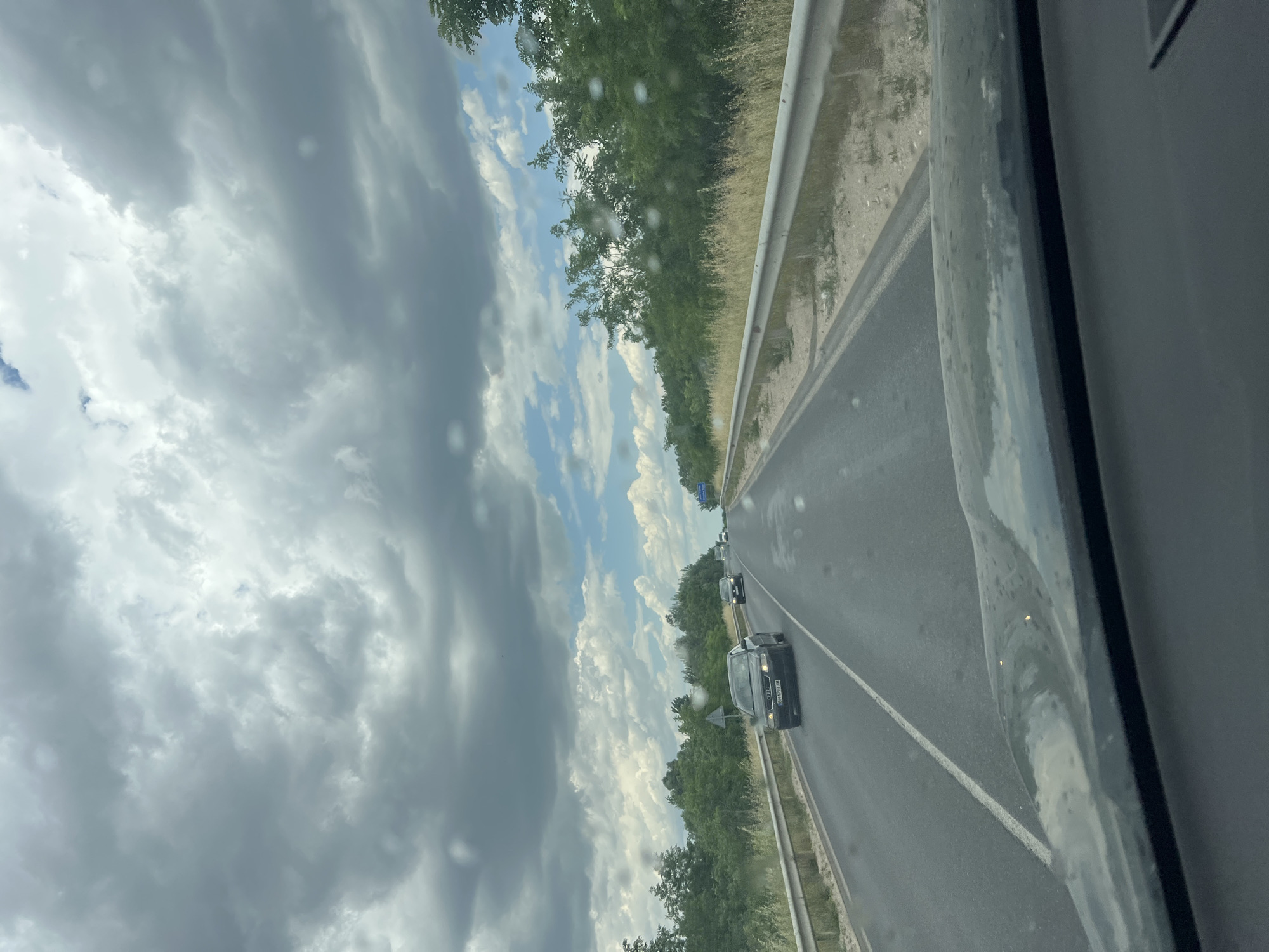 The image depicts a view from inside a vehicle traveling on a multi-lane highway in Kalekovets, Bulgaria. The highway is lined with trees and has a guardrail on the right. Several vehicles, including a dark-colored sedan, are visible on the road ahead. The sky is cloudy, with patches of blue visible. The inside of the vehicle includes a window frame with water droplets.