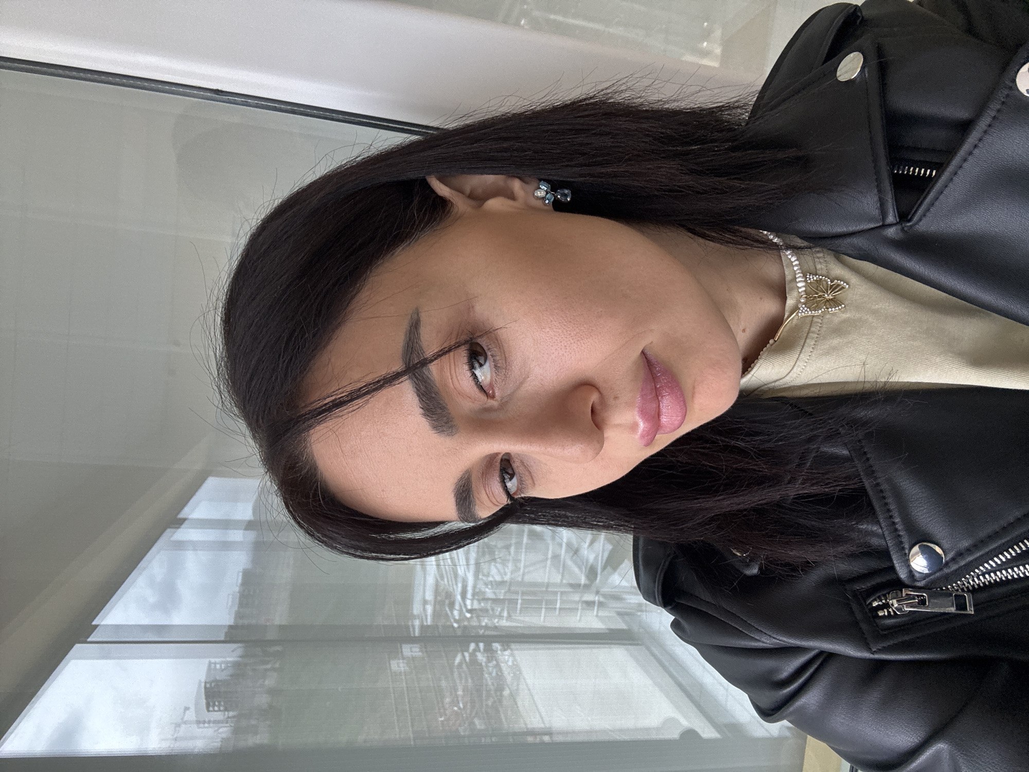A female subject is depicted in a close-up, chest-up self-portrait indoors, positioned in front of a large window. She has dark hair, styled with a side part, and is wearing a black faux leather motorcycle jacket over a light beige top. A gold-colored butterfly pendant necklace with small stones is visible around her neck, and a small, multi-gemstone earring, primarily blue/teal, is present in her right ear. Her lips are lightly colored with a pink hue, and she maintains direct eye contact with the camera. The background, visible through the window, shows an overcast sky and several modern high-rise buildings, some of which appear to be under construction with a visible crane. This urban view, characterized by contemporary development and high-rise architecture, is consistent with the **Saburtalo District** of Tbilisi, Georgia, suggesting the subject is located in a high-floor apartment or office in that area.