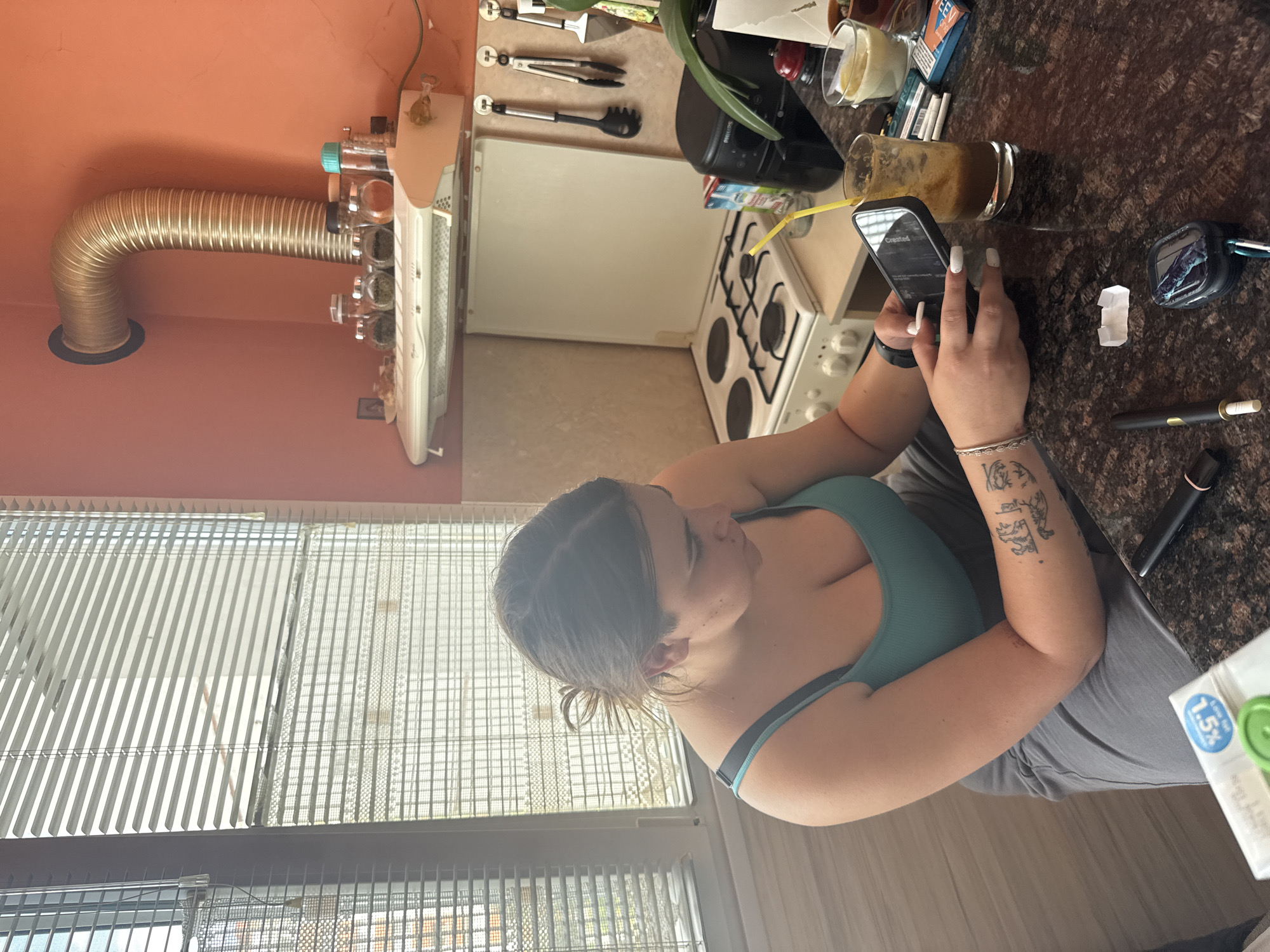 The image captures a candid moment of a woman seated in a kitchen in Kyustendil, Bulgaria, during daytime, indicated by bright light filtering through vertical window blinds. The woman, with her dark hair pulled back, is wearing a teal tank top and gray pants. She is engrossed in her smartphone, which she holds in both hands, revealing long white manicured nails and a smartwatch on her left wrist. Tattoos are visible on her left arm.
The kitchen features an orange-colored wall with a metallic corrugated vent pipe extending from it, likely for an extractor fan. Below this, a wall-mounted spice rack holds several glass spice jars. A white gas stove with four burners is partially visible on a countertop made of dark, speckled granite. Other items on the counter include a Philips coffee machine, various kitchen utensils hanging on the wall, a glass containing a light-colored drink, another glass with a dark brown beverage and a yellow straw, an earbud case, a vape pen, a pack of "EET" cigarettes, and a small white rectangular object. The smartphone screen faintly displays "Created for". A white carton marked "1.5%" is visible on the floor beside her. The scene suggests a relaxed, domestic setting.