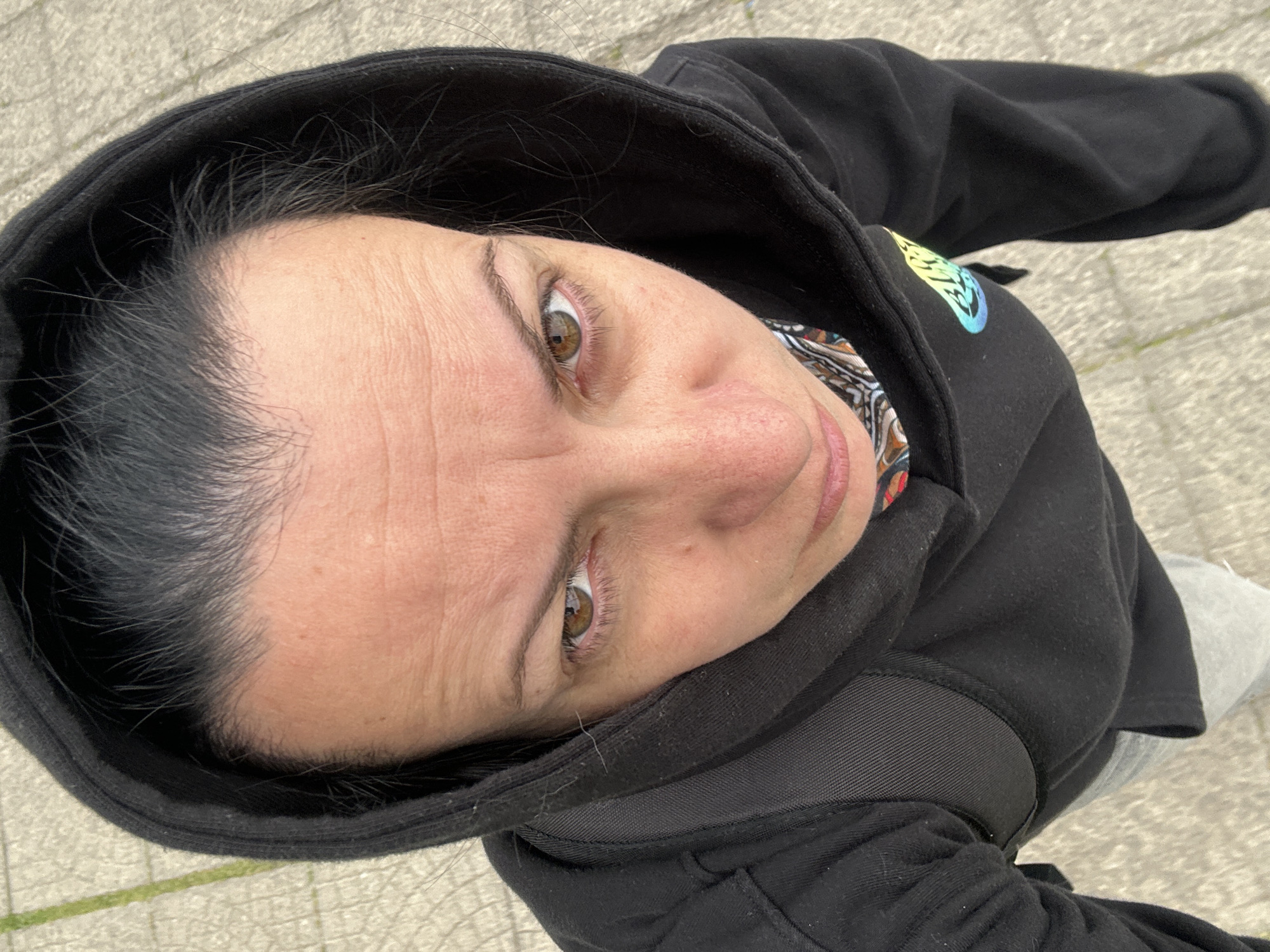 A person with fair skin and brown eyes is looking up at the camera from a low angle. They are wearing a black hooded sweatshirt, the hood pulled up around their face. The sweatshirt appears to have a colorful emblem on the upper left shoulder. The person's hair is dark, and some strands are visible around the edges of the hood. The background shows a textured surface, possibly pavement or stone tiles. Given the lack of distinct architectural features or other identifying landmarks, it is not possible to confirm the location as Sofia, Bulgaria, from this image alone.