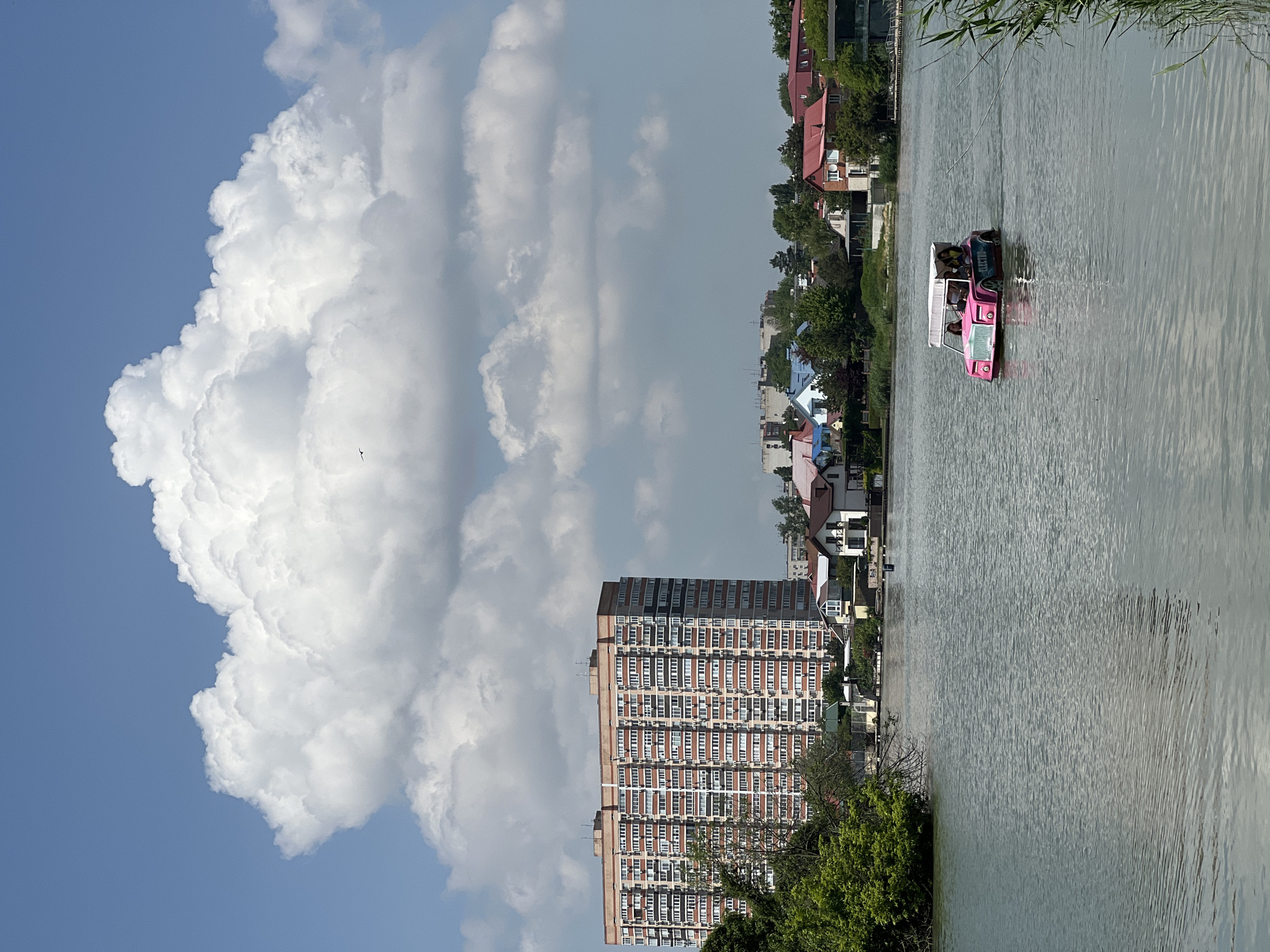A medium, landscape-oriented shot portrays a scene on the Kuban River in Krasnodar, Russia.
The sky, predominantly blue, features large, puffy cumulus clouds, contrasting with the tall apartment building situated near the river's edge. The building has a repetitive pattern of windows and balconies.
A pink "electric" boat with a white canopy, carries several occupants on the river. The river reflects the sky and surrounding buildings. Trees and green vegetation line the riverbank, with residential houses nearby.
The scene appears to be captured on a clear day.