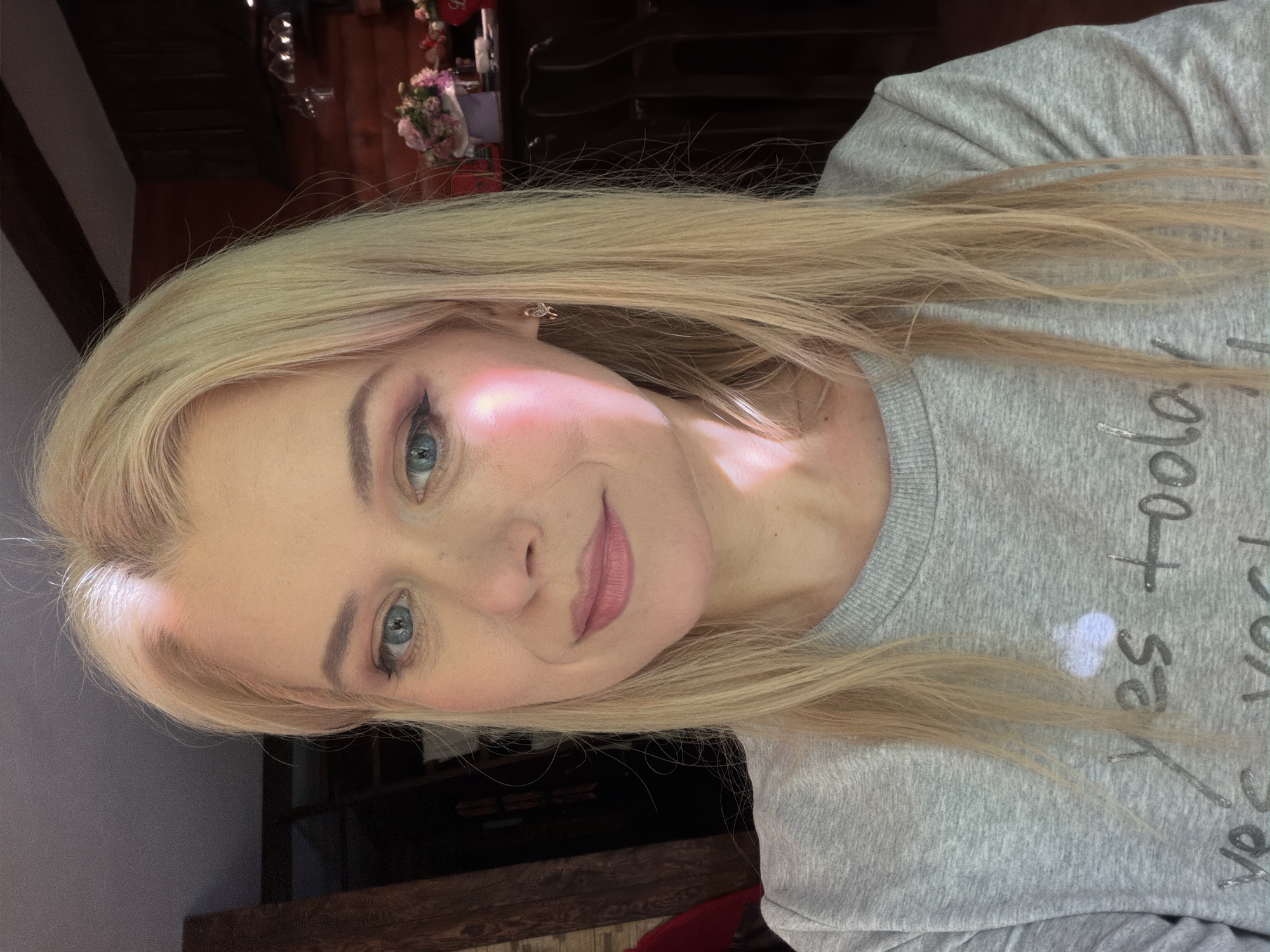 The image is a close-up selfie of a young blonde woman. She has blue eyes, fair skin with a light blush on her cheeks, and is wearing light pink lipstick. Her hair is long and blonde, framing her face. She is wearing a light gray sweatshirt with text on it. The text appears to be in a stylized font and some of the letters are visible, including "yes" and what looks like "today". The lighting in the image suggests it could be daytime, with some light shining on her face. The background is out of focus but appears to be indoors, with a dark wooden surface and some floral decorations visible. The overall mood of the image is relaxed and friendly. The location context is Sochi, Russia.