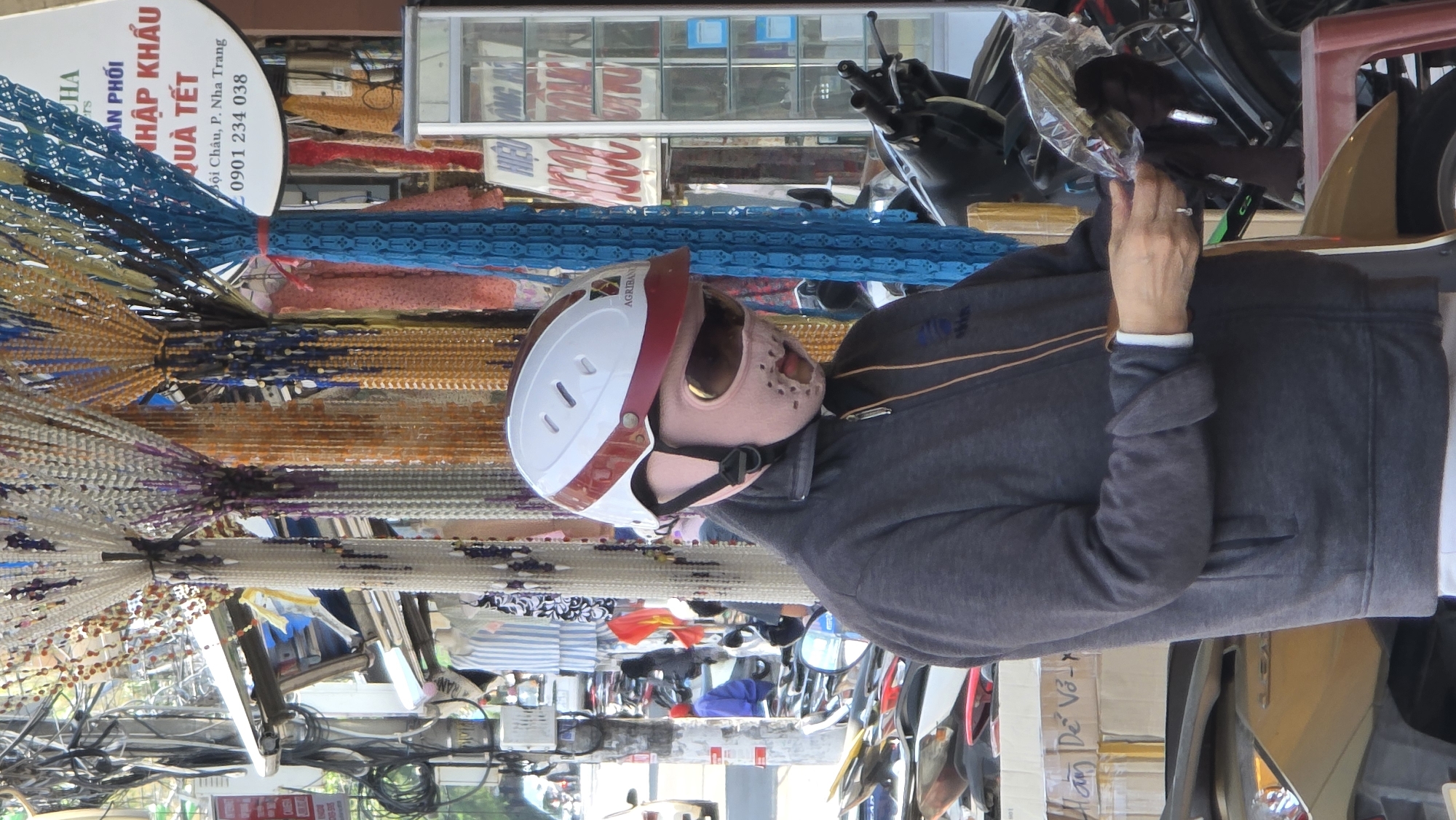 A person wearing a motorcycle helmet and a full-face mask is seen outdoors in a street scene. The individual is wearing a gray jacket with a white collared shirt underneath and has a ring on their finger. They appear to be examining something held in their hands.
Behind the person, multiple strands of beads in varying colors (white, orange, purple, blue, and black) hang vertically. Further back, there are parked motorcycles and scooters, reflecting the density of traffic often seen in urban Asian environments. There are also boxes with Vietnamese writing on them.
A sign in the background has Vietnamese text: "Hàm Dể Vỏ -", "ÂN PHỐI NHẬP KHẨU QUÀ TẾT Bội Châu, P. Nha Trang 0901 234 038", indicating that this is in Nha Trang, Vietnam, specifically in the Bội Châu ward. The sign also mentions "Quà Tết" (Lunar New Year gifts), suggesting this is a business selling or displaying items related to the Tết holiday.