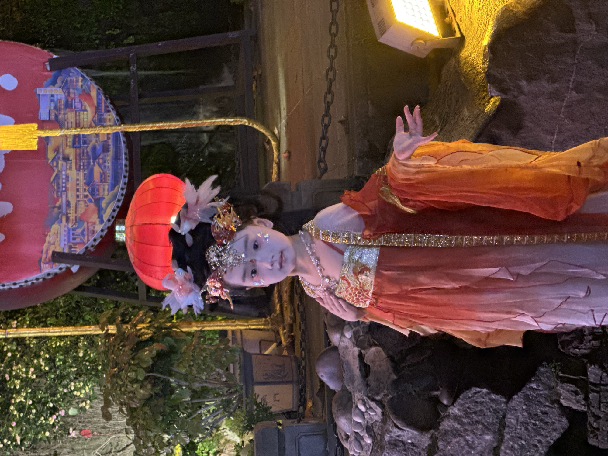 The image depicts a young girl dressed in elaborate traditional Chinese-style attire, posing during the evening or night. She stands centrally, looking directly at the camera with a calm expression, her hands held gracefully. Her costume features a multi-layered dress in shades of orange, red, and cream, embellished with golden embroidery and sequins. She wears an ornate headpiece adorned with flowers and a crown, a delicate necklace, and bracelets. Glitter is visible on her face, adding to the festive appearance.
The setting is an illuminated outdoor or semi-outdoor space, possibly a cultural garden or exhibit. A bright yellow spotlight positioned in the upper right casts light on the scene. Behind the girl, to her left, a large red Chinese lantern with intricate patterns is visible, alongside a decorative fan or screen painted with a detailed cityscape of traditional Chinese architecture in blue, yellow, and red. Another red lantern is partially seen near her head. The background also includes dark metal chains, rocks, and green foliage with small, light-colored flowers. The ground around her is composed of various rock formations.
The scene suggests a cultural event, performance, or themed photoshoot, likely taking place in a location like Yuzhong District, China, given the prominent traditional Chinese elements. No legible text is visible in the image.