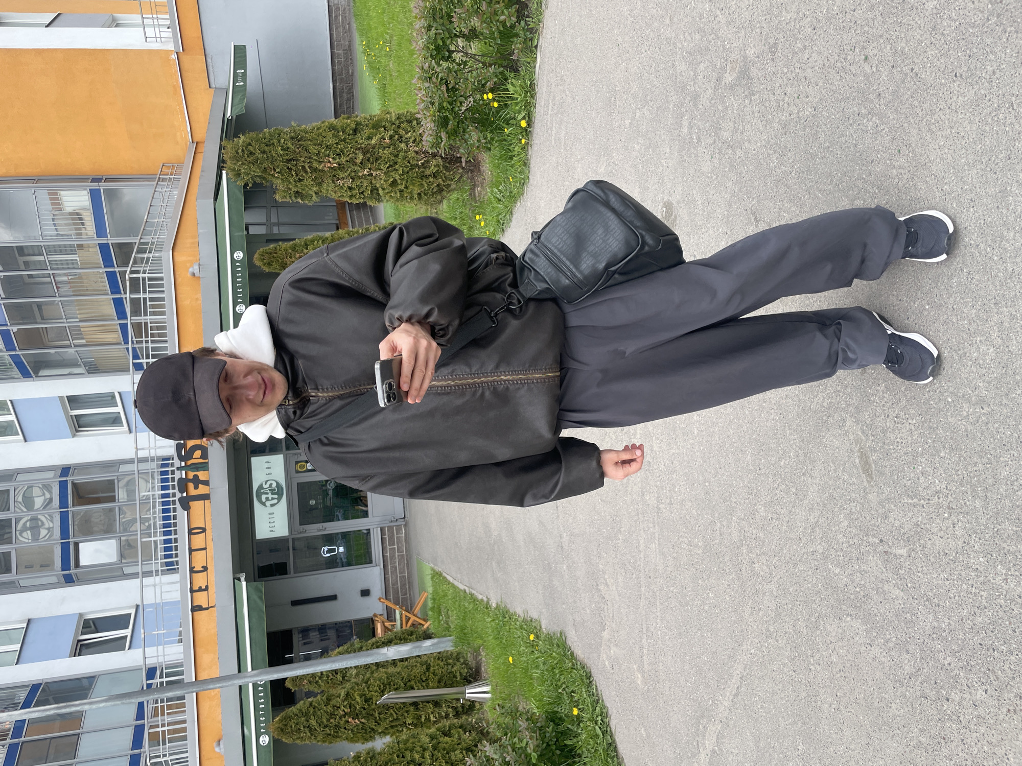 A person in a black jacket and pants stands on a sidewalk in front of a building. The building is labeled "PECTOPAH 17.05", which is in Cyrillic. The person is holding a cell phone in their left hand, and a black bag is slung across their torso. Grass and bushes are visible alongside the sidewalk and in front of the building. The building appears to be multi-story with numerous windows. The location is in the city of Unknown, Russia.