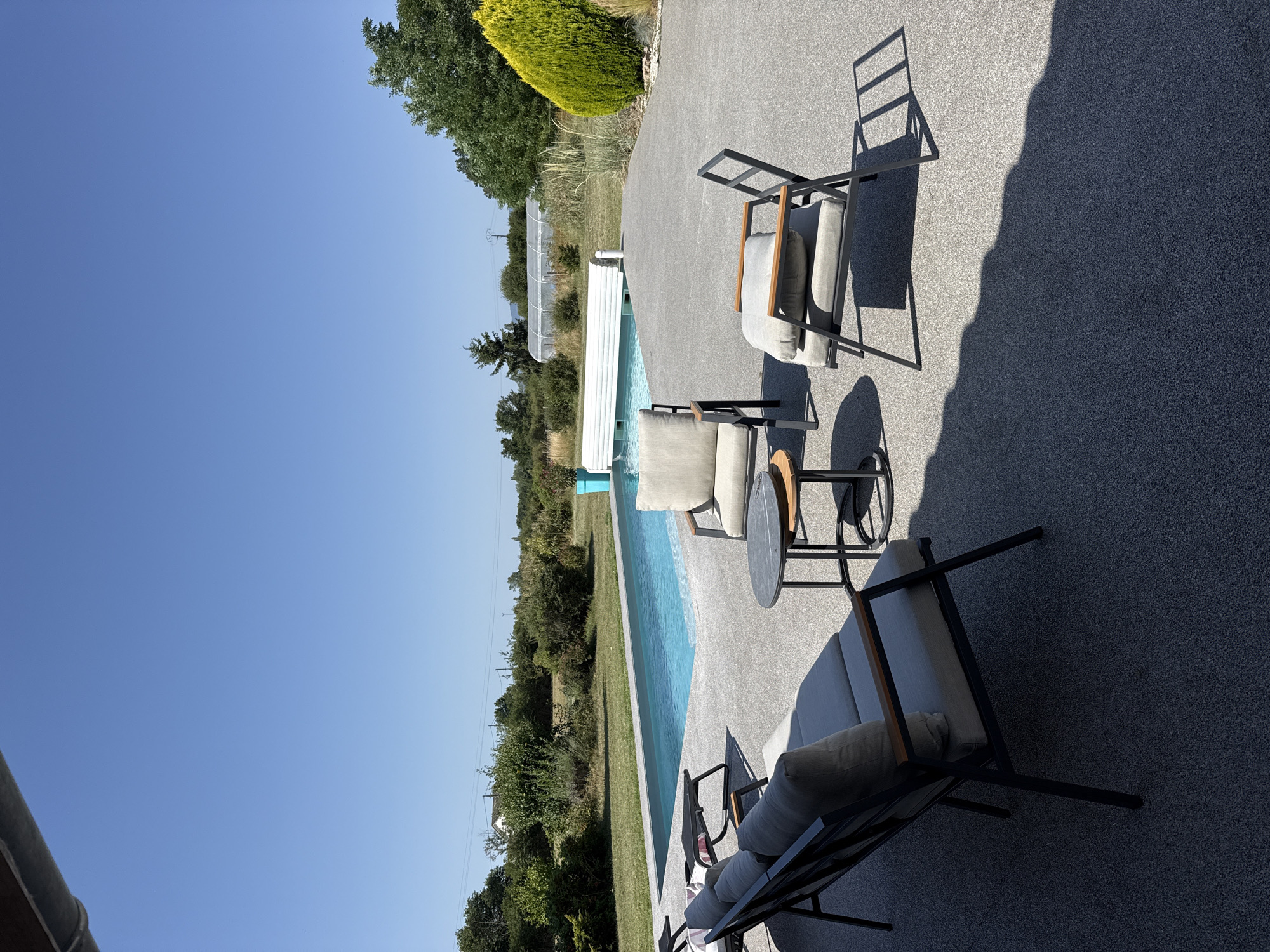 This is a medium shot, eye-level outdoor photograph taken on a clear, sunny day. The scene is a residential backyard in Veauchette, France, featuring a swimming pool and patio furniture.
In the foreground, a grey stone patio is visible. On the left side of the patio, a dark metal sofa with grey cushions is positioned. To the right of the sofa, there are two dark metal chairs with grey cushions and wooden armrests. A small, round, dark grey metal table with a wooden top sits between the chairs.
The swimming pool, which is rectangular and appears to be in-ground, occupies the middle ground of the image. The water is a bright blue. A white pool cover is partially rolled out over the pool.
In the background, a lawn with green grass is visible, bordered by various trees and shrubs. Beyond the lawn, a greenhouse structure with a white cover can be seen. A single-story house is partially visible in the far distance. The sky above is a clear, bright blue with no clouds. The overall impression is of a well-maintained outdoor living space.