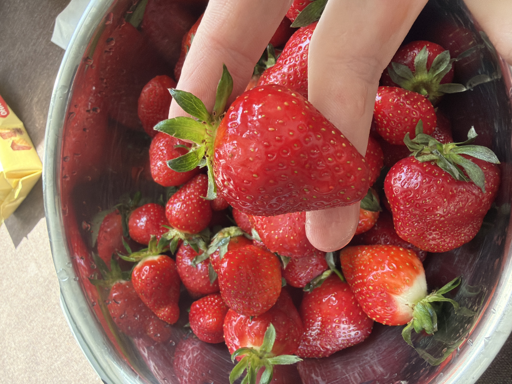 A close-up, high-angle shot shows a stainless steel bowl filled with fresh red strawberries. A human hand with pale skin is partially visible, holding a large strawberry with its green leaves. The bowl is filled with a generous amount of strawberries. A partially visible yellow packaging is in the background on the left. The scene is well-lit with natural light. Location information is not available.