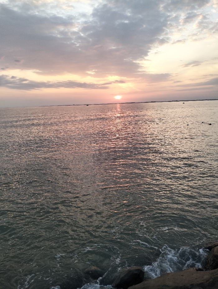 The image depicts a tranquil seascape at sunset. A wide expanse of water, reflecting the sun's golden glow, fills the frame. Gentle waves ripple across the surface, meeting rocky terrain at the bottom. The sky is adorned with soft, muted clouds, tinged with hints of pink and gray. The sun, appearing as a small, bright orb on the horizon, casts a shimmering path across the water. The scene conveys a sense of peace and serenity.